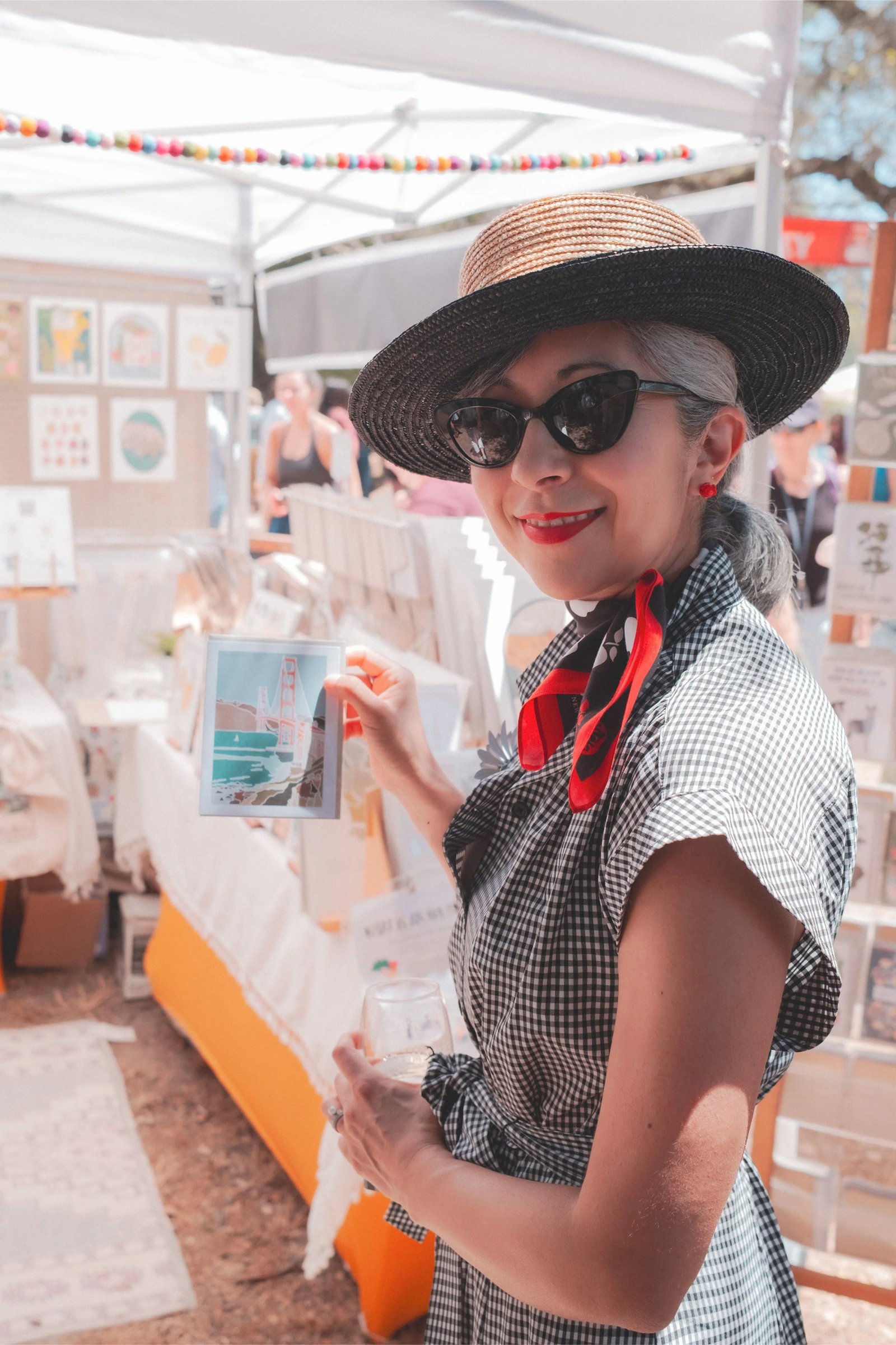 A smiling woman in a hat and sunglasses holds a framed illustration of the Golden Gate Bridge at an outdoor market.