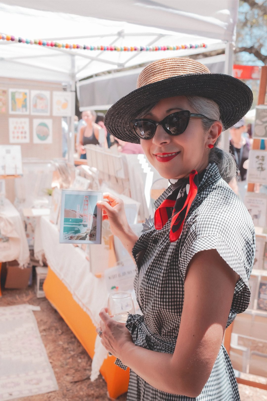 A smiling woman in a hat and sunglasses holds a framed illustration of the Golden Gate Bridge at an outdoor market.