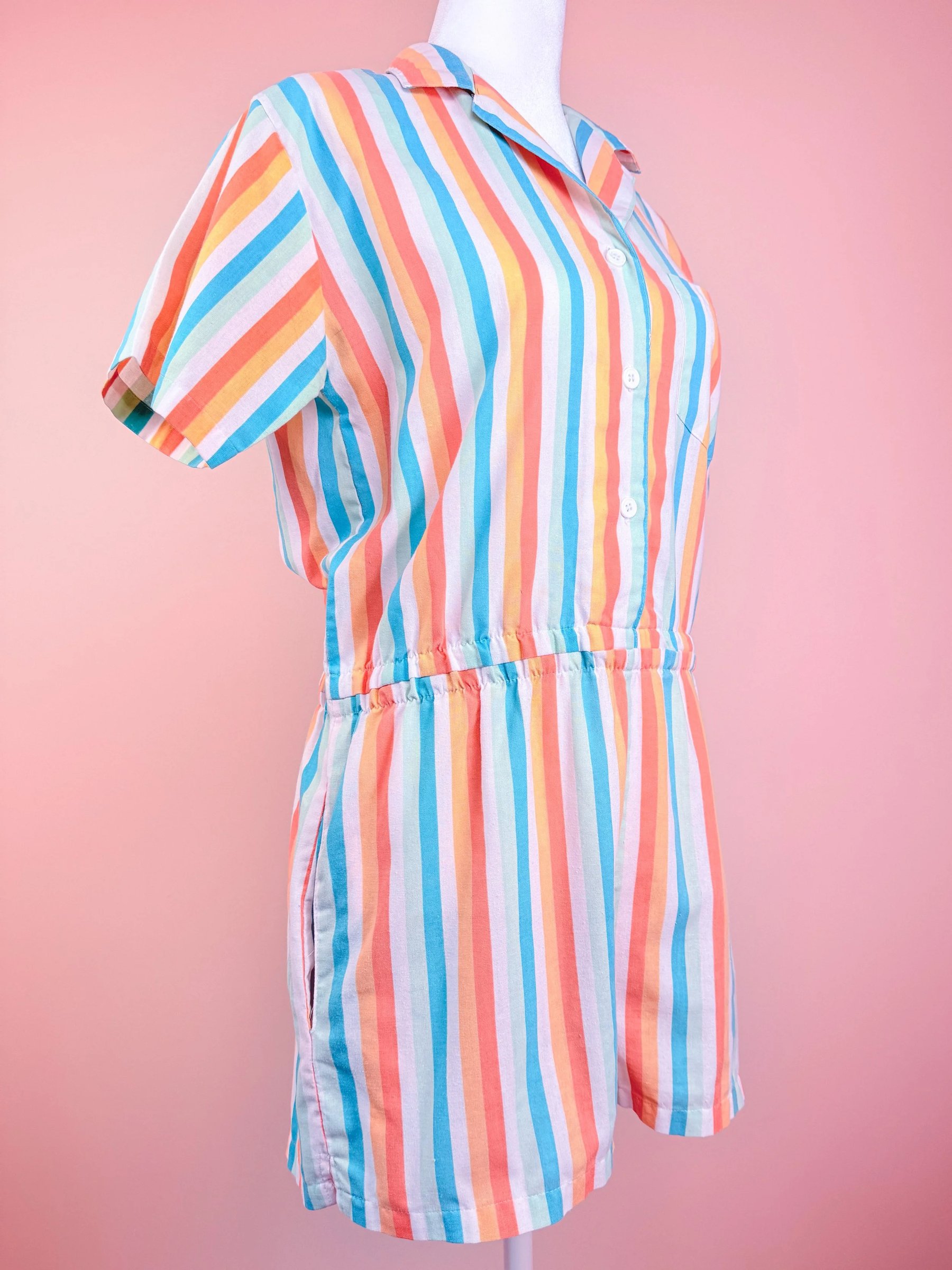 A colorful striped jumpsuit featuring short sleeves and an elastic waistband against a pink background.