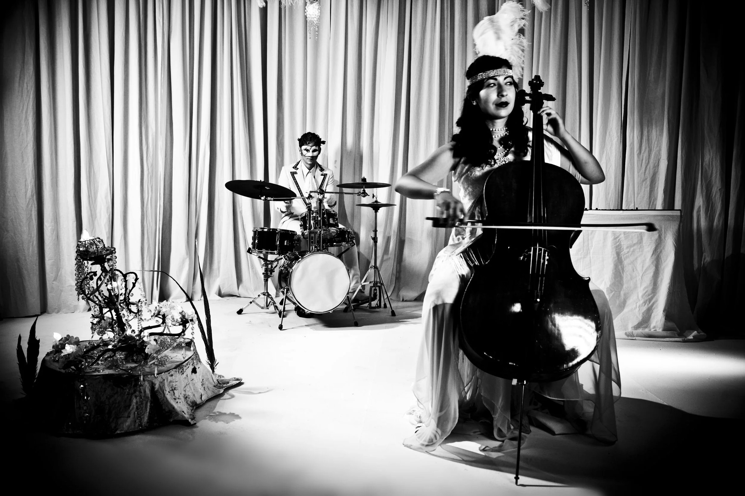 A woman in a white gown plays the cello while a drummer is positioned in the background, surrounded by draped fabric and floral arrangements.