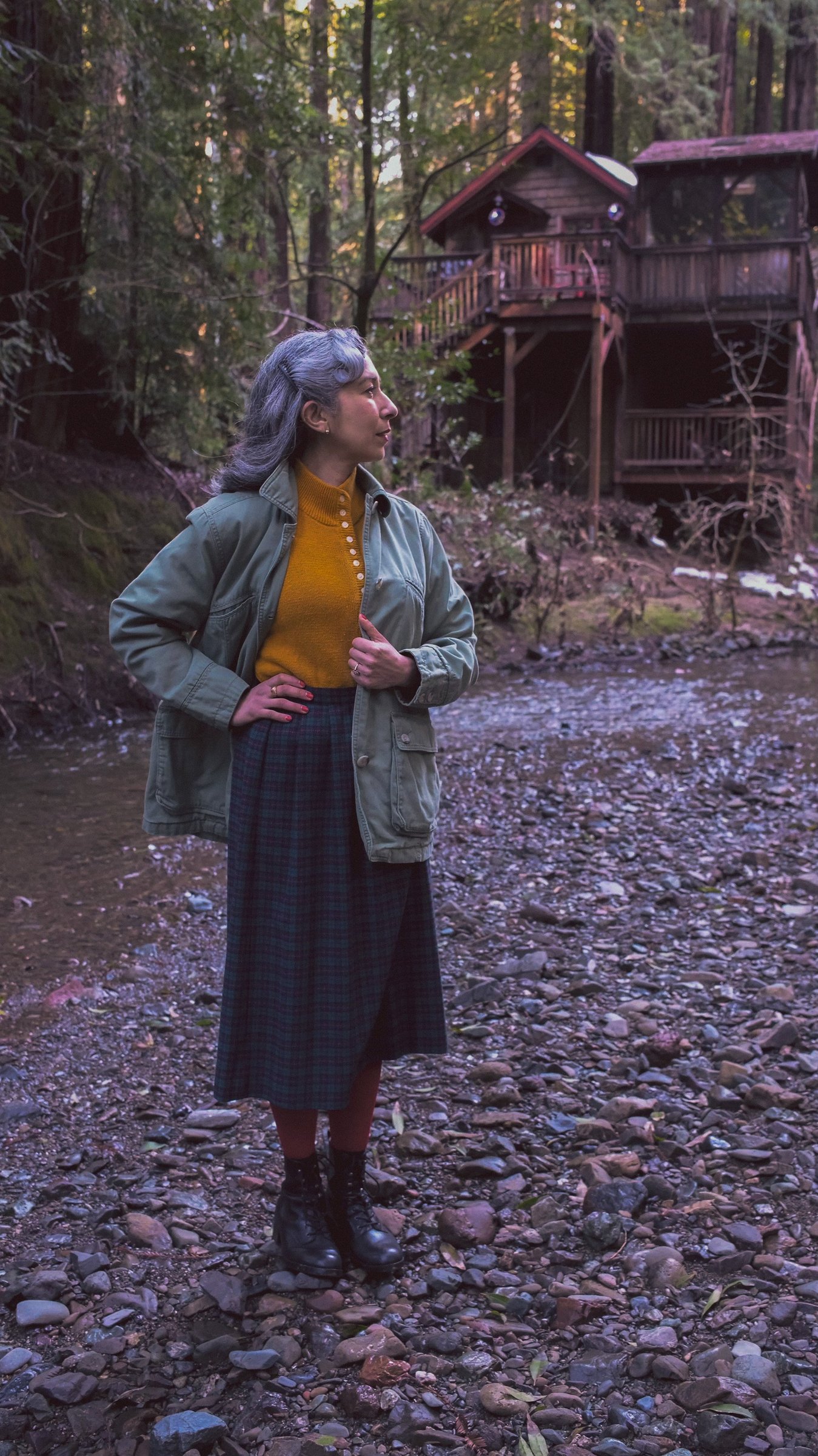 A woman with gray hair wears a green jacket, orange sweater, and plaid skirt while standing on rocky ground near a wooden cabin in a forested area.