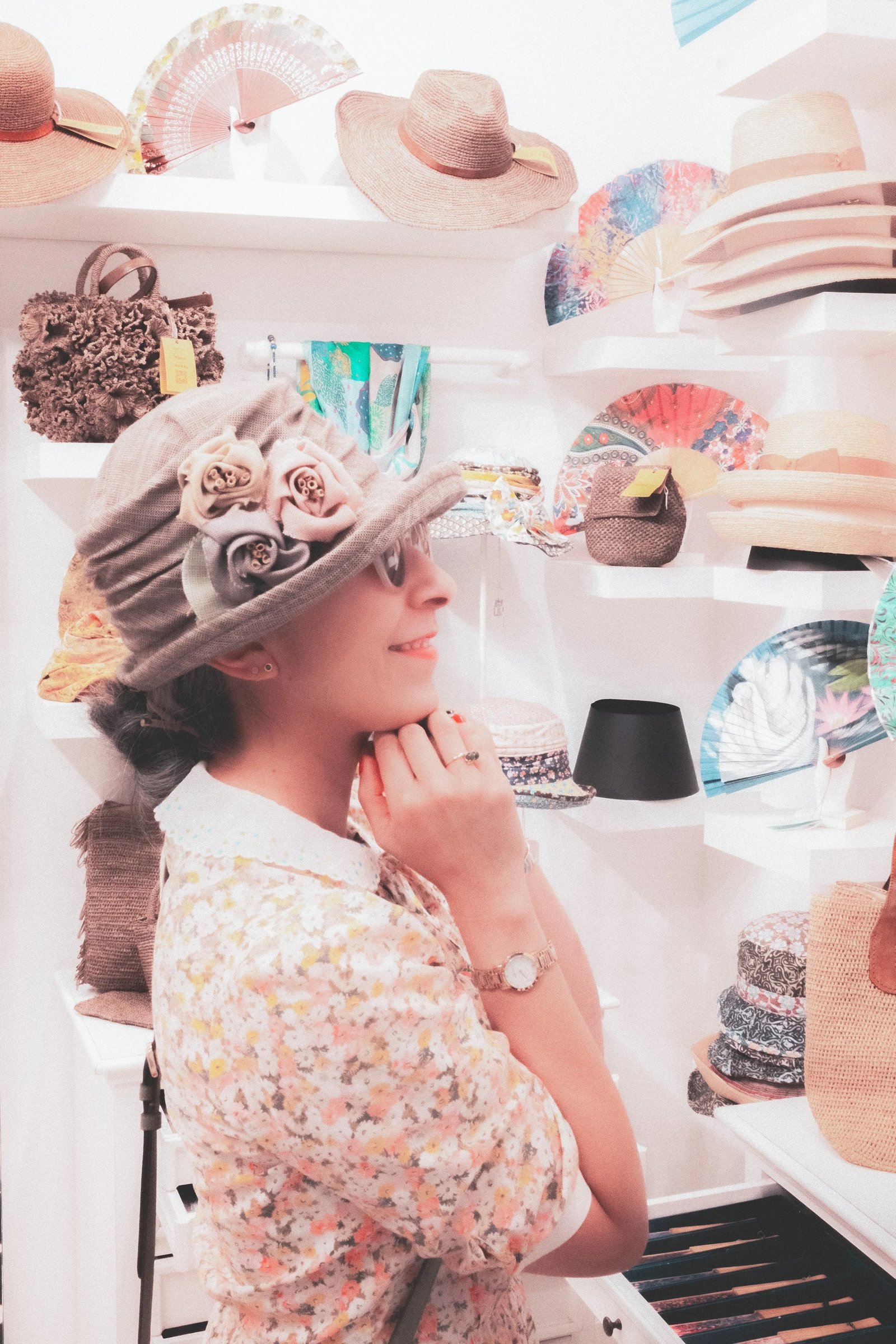 A woman wearing a floral blouse and a decorative hat smiles while browsing a variety of hats and fans in a store.
