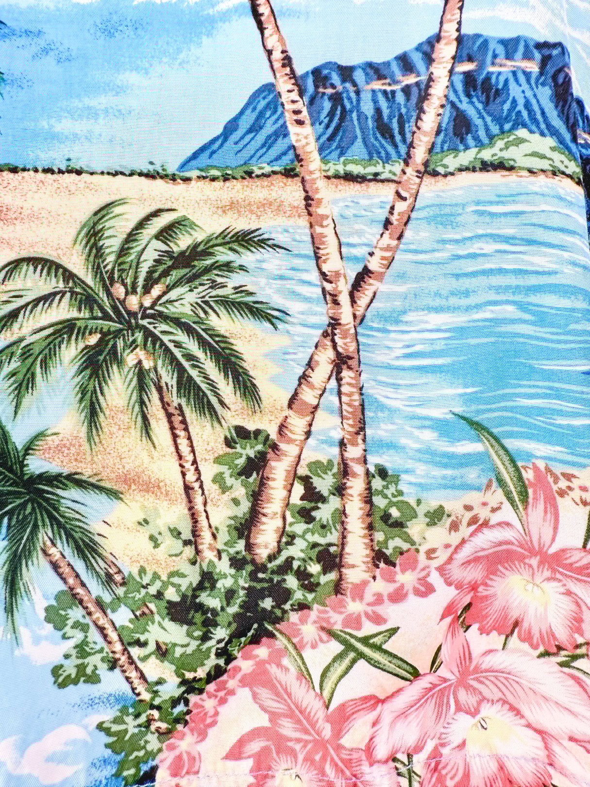 The Aloha Darling Top features a vibrant blue fabric with a 1960s tropical print, showcasing palm trees, hibiscus flowers, and a scenic beach landscape.