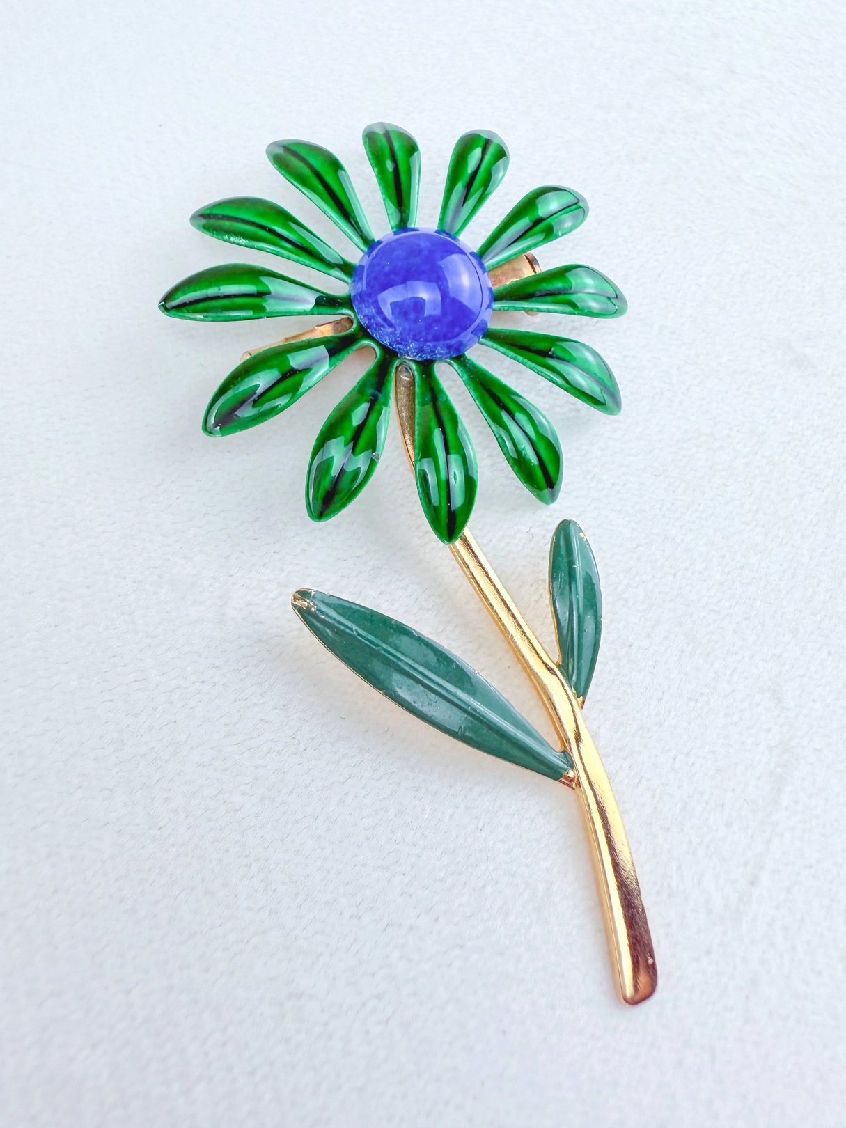 Vintage-style floral brooch featuring green enamel petals, a blue center gemstone, and gold-tone leaf and stem accents.