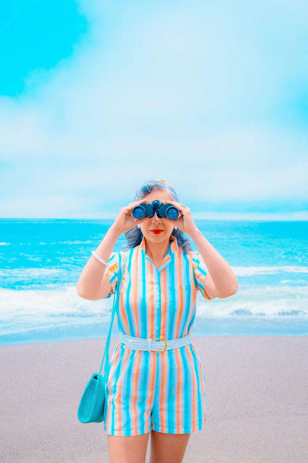 A woman in a striped shirt holds binoculars while standing on a beach with ocean waves in the background.