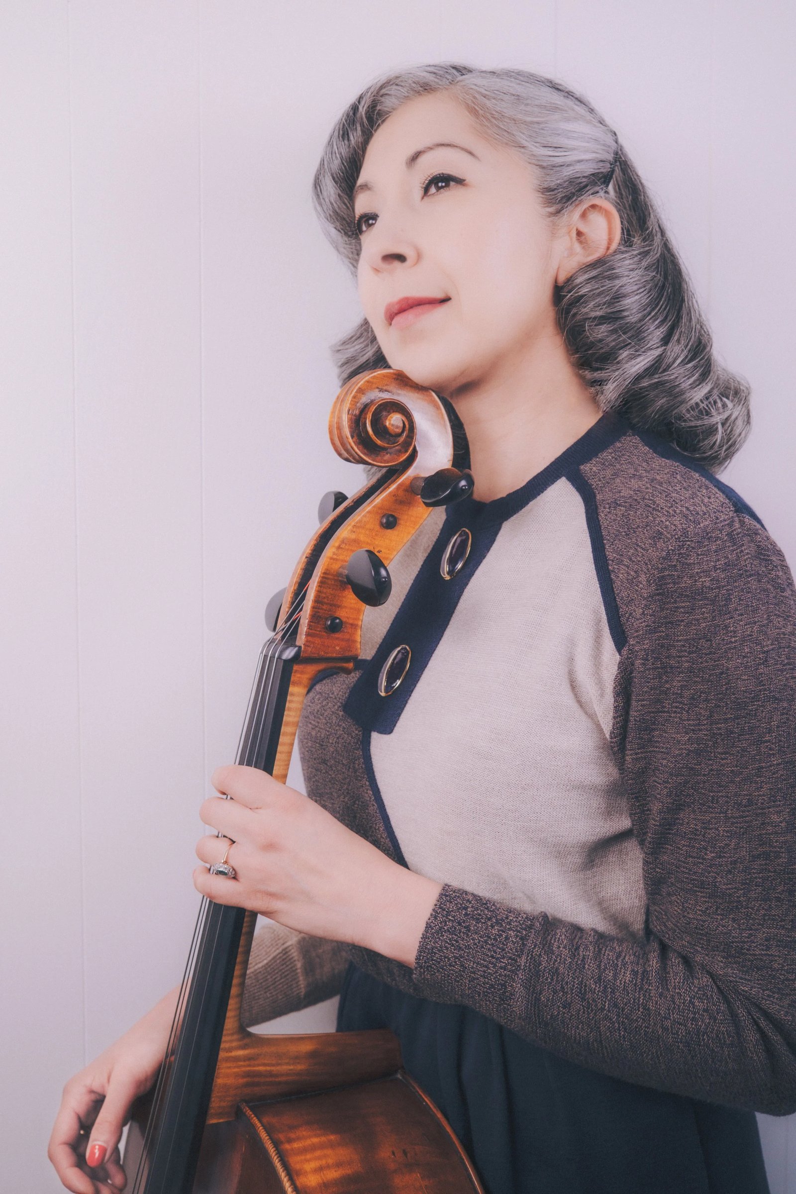 A woman with silver hair holds a cello, wearing a vintage-inspired sweater with a navy and beige design.