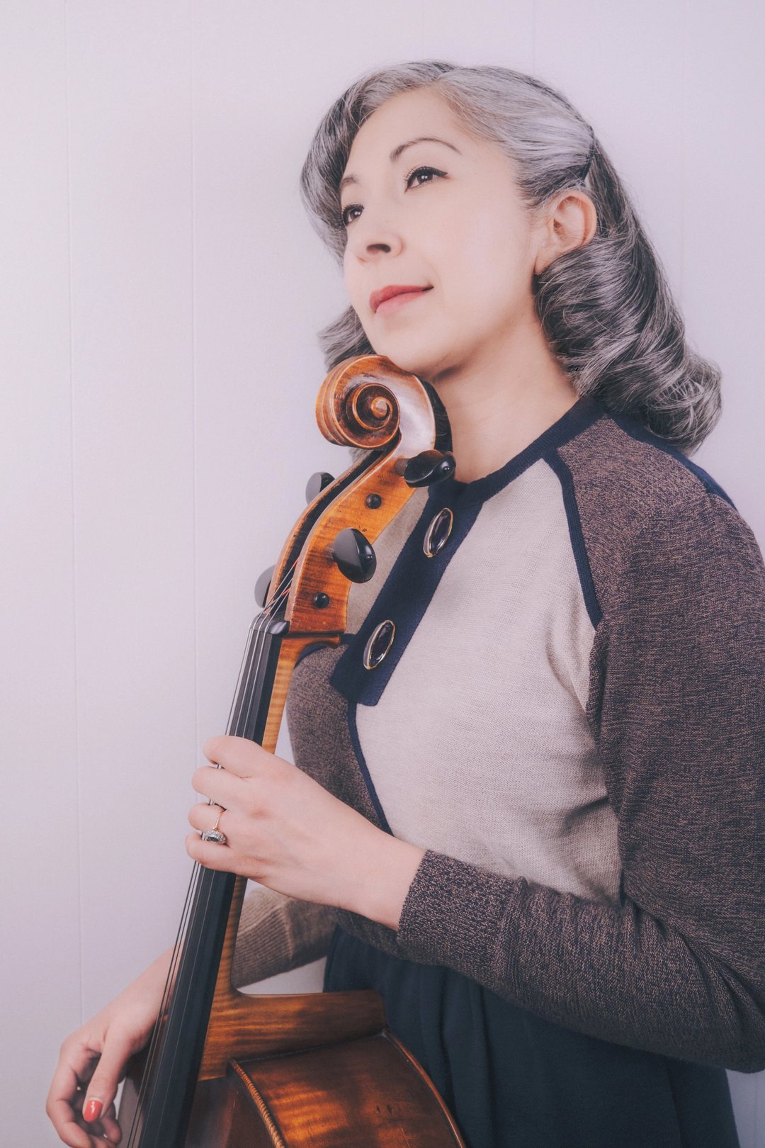 A woman with silver hair holds a cello, wearing a vintage-inspired sweater with a navy and beige design.