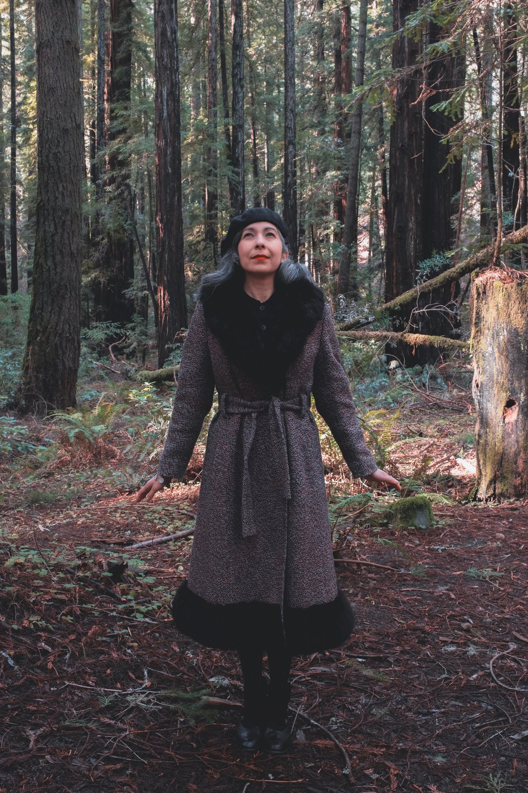 A person wearing a dark coat stands among tall trees in a forest, with their arms raised and a serene expression.