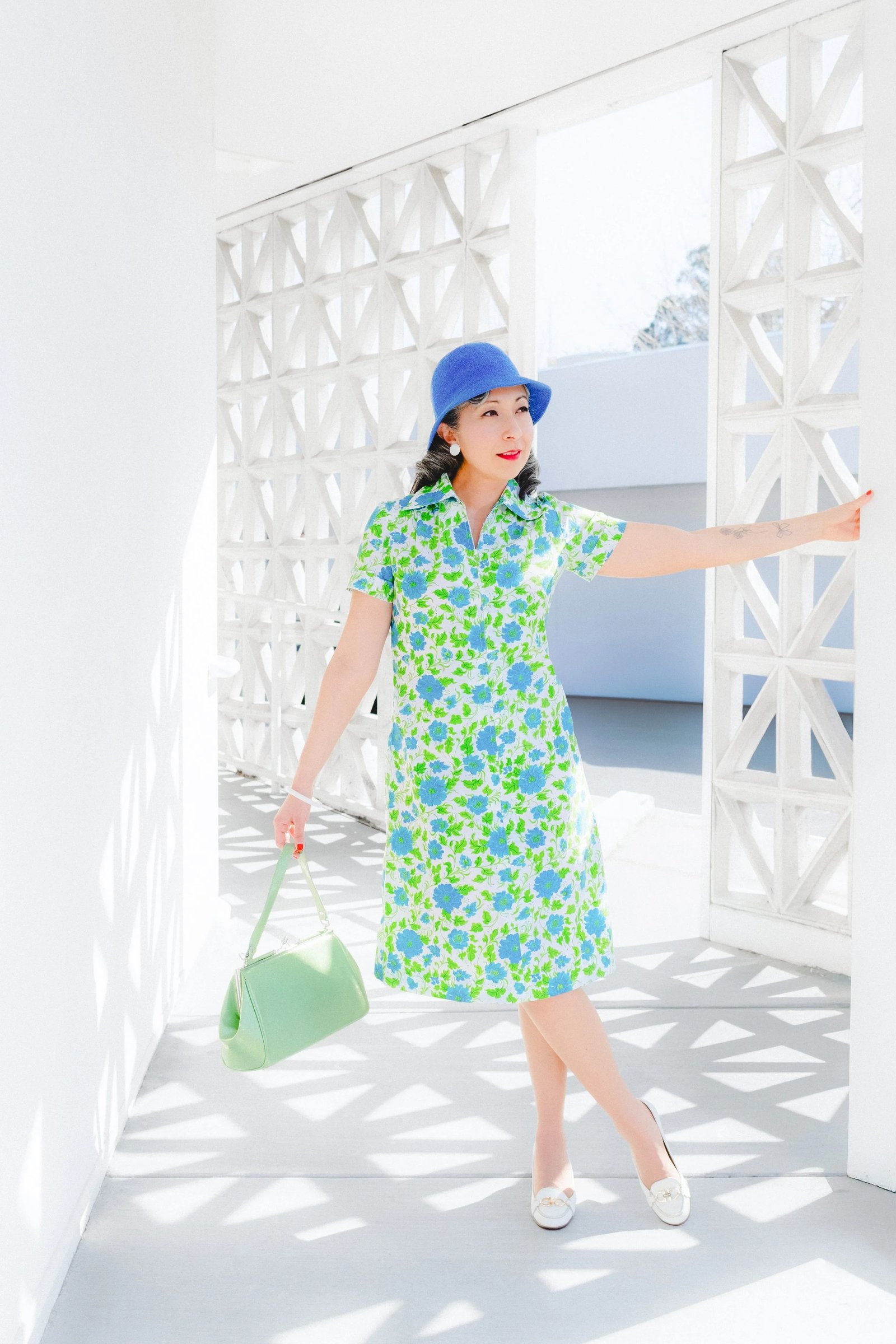 A woman poses in a green floral dress and blue hat, holding a clear handbag, against a bright, white backdrop.