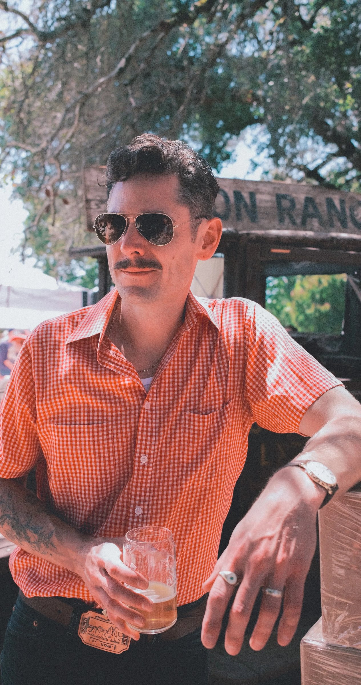 A man in a checkered orange shirt and sunglasses holds a glass of beer, leaning against a wooden bar in a sunny outdoor setting.