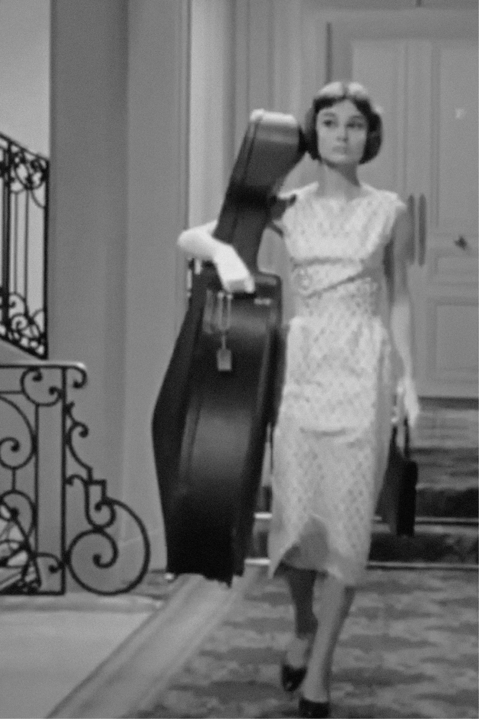 A woman in a vintage dress walks through a doorway carrying a guitar case and a small handbag.