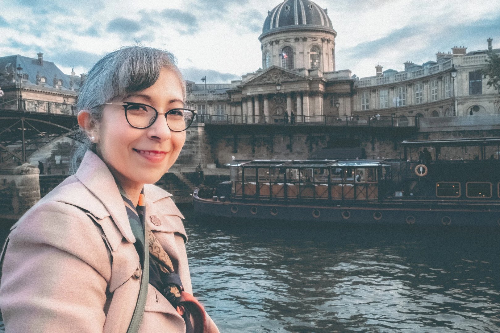 A woman with glasses smiles while sitting by a river with a historic building in the background.