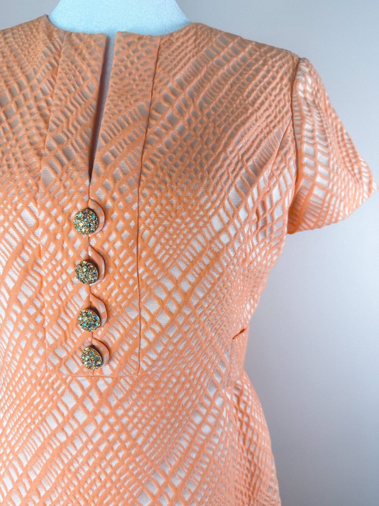The Orange Jubilee Dress features a textured orange fabric, short sleeves, and four decorative buttons with a sparkling design, reflecting 1960s fashion trends.