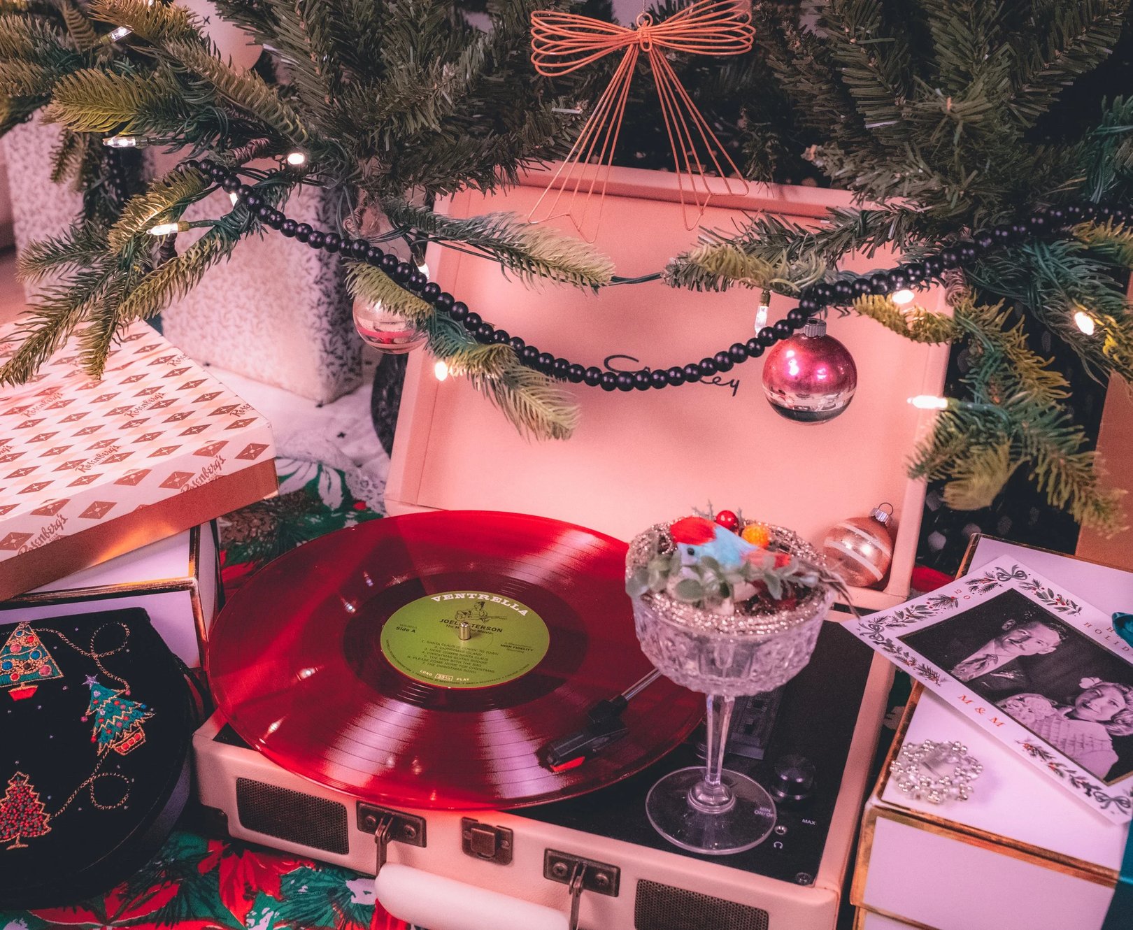 A vintage record player with a red vinyl record is surrounded by holiday decorations, including a decorated Christmas tree and festive ornaments.