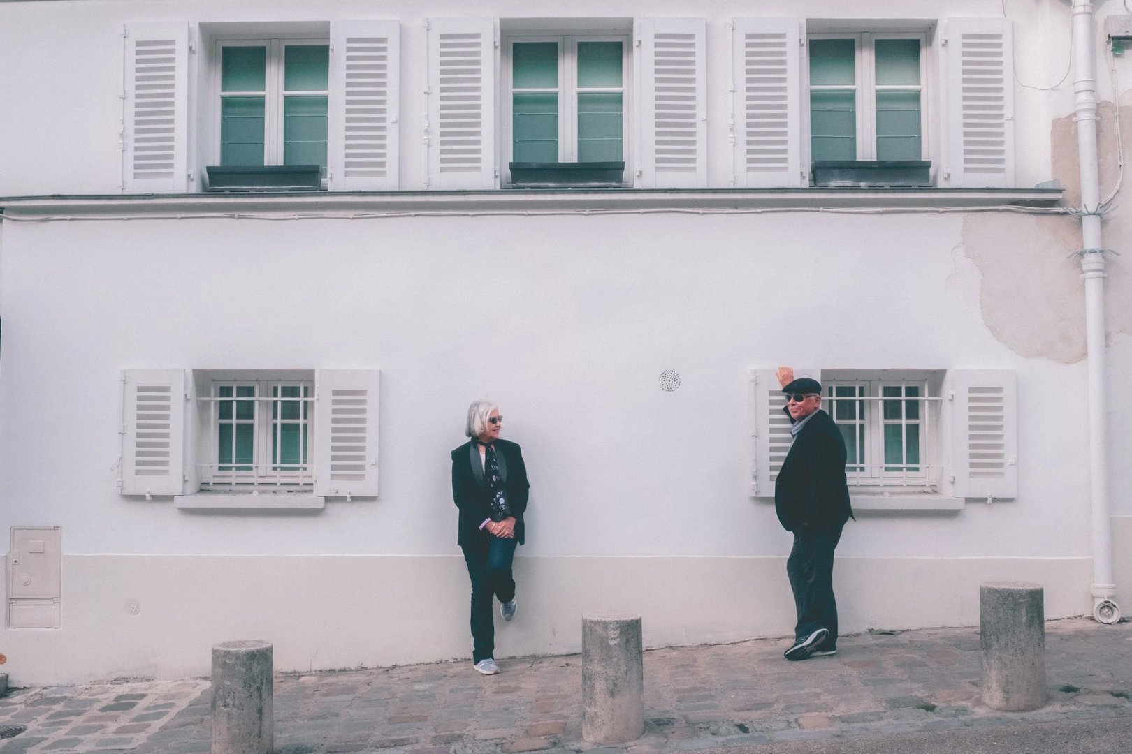 Two individuals stand beside a white wall with windows, dressed in black, against a light backdrop.