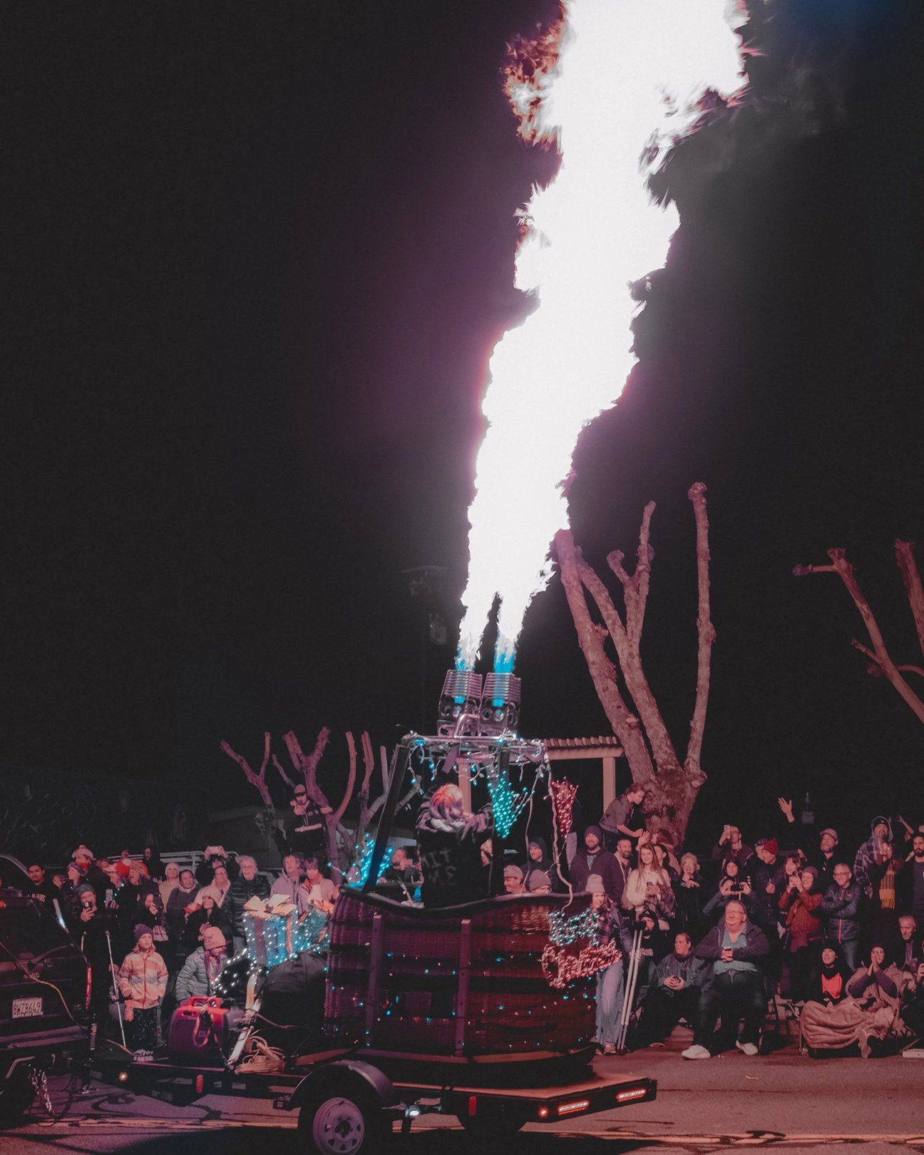 A crowd watches as a colorful fire-breathing robotic sculpture emits large flames at night.