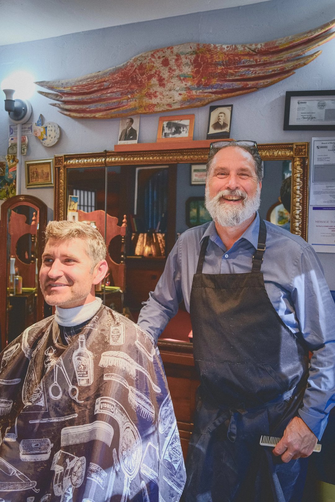 A barber stands next to a seated customer in a vintage barbershop, both smiling while surrounded by barber tools and photographs.