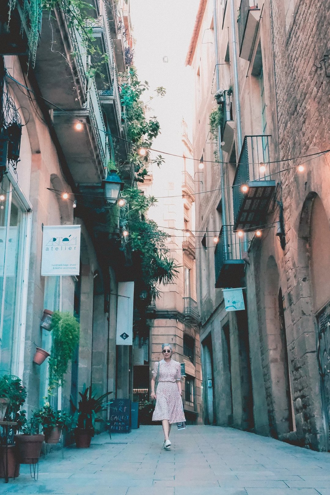 A woman in a sundress walks down a narrow, plant-lined alleyway adorned with string lights and shop signs.