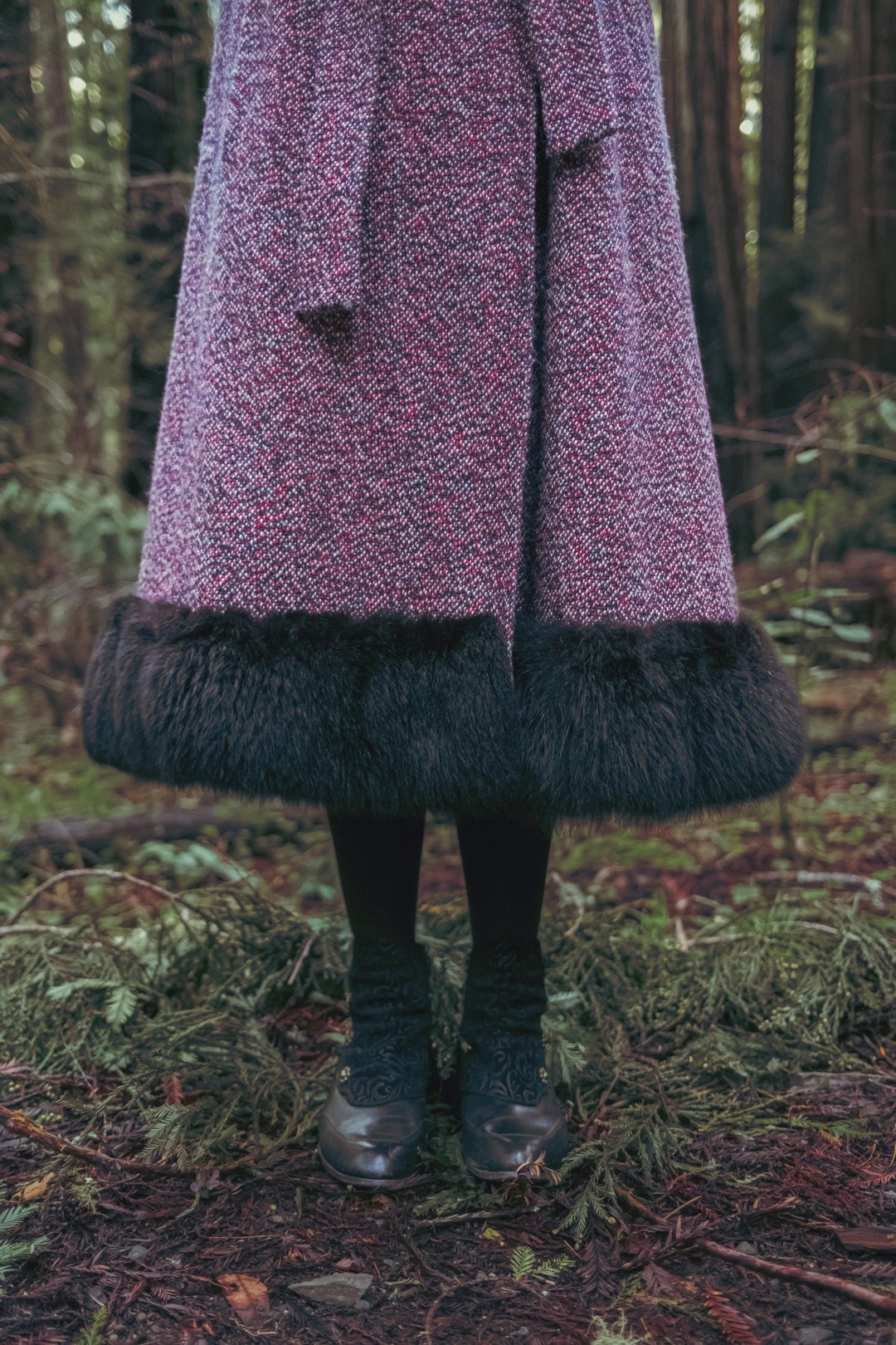 A person’s legs are visible in a forest, wearing a long purple and pink herringbone coat with a fur hem and black shoes.