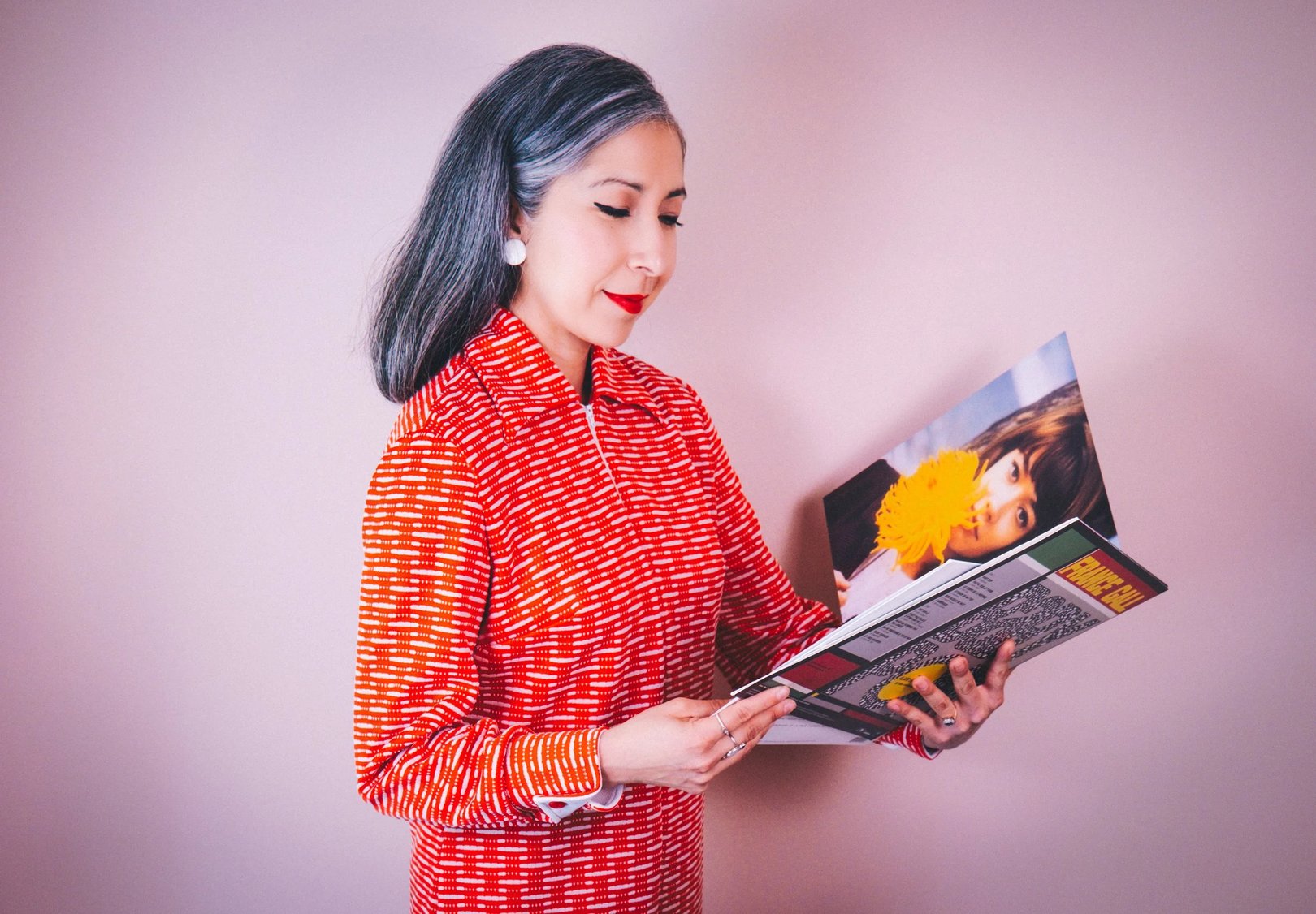 A woman with gray hair wearing a red patterned blouse holds a magazine while standing against a pink background.