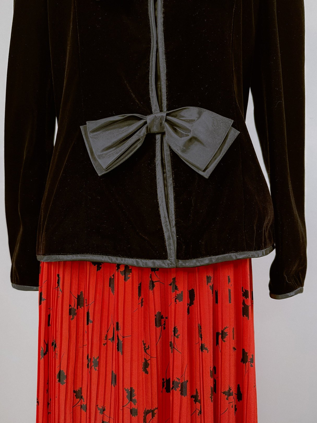 Black velvet women's jacket from the 1980s featuring a bow detail at the waist, worn over a red pleated skirt with a floral pattern.