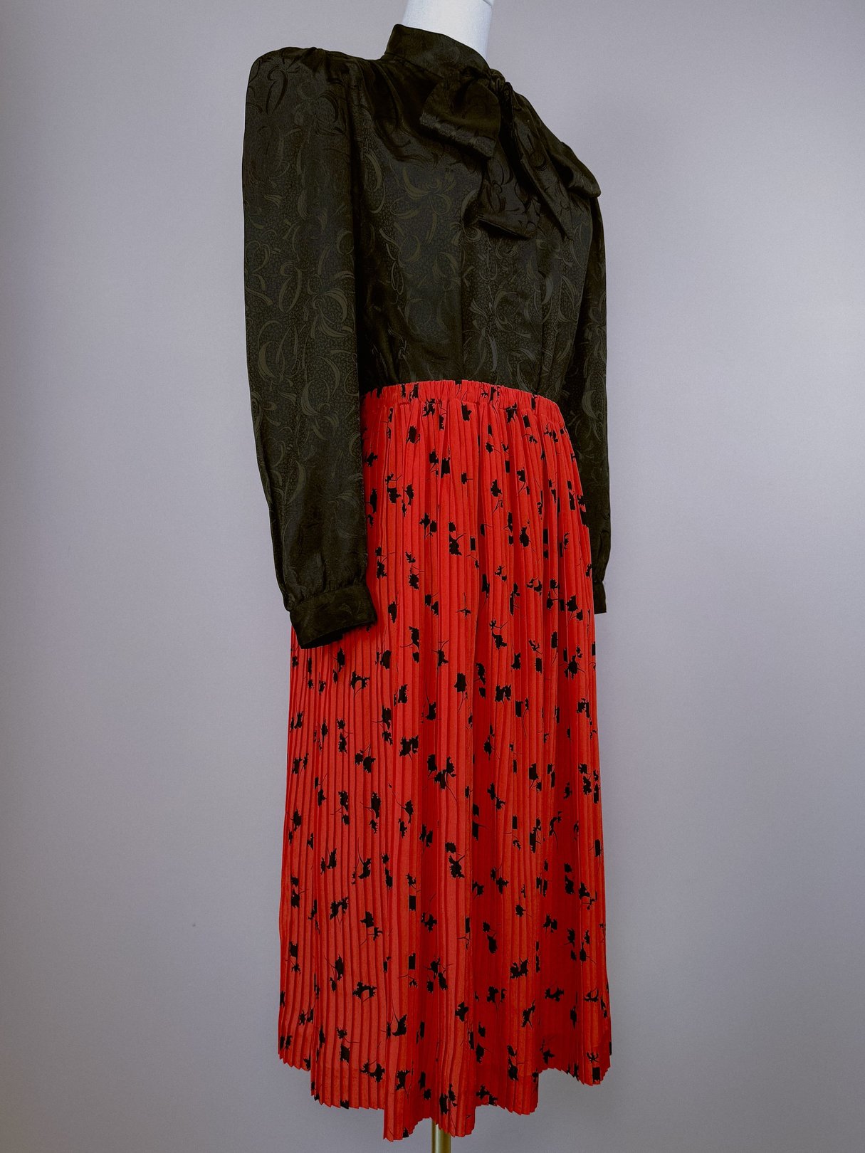 1970s women's pleated skirt in red with black floral print, paired with a dark green, textured blouse featuring a bow tie.