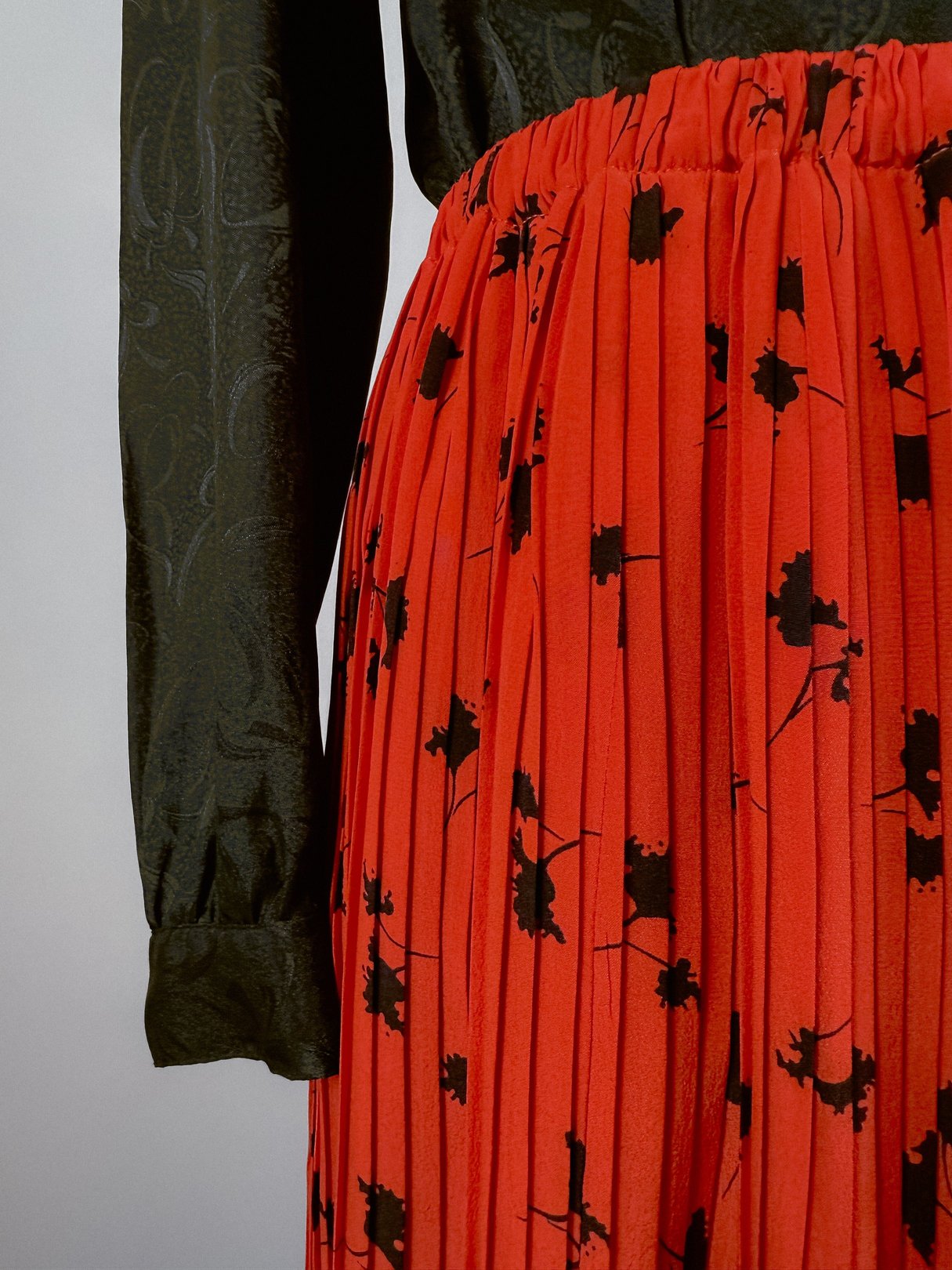 A pleated skirt in red with a floral pattern, designed for women, featuring a high waistband and paired with a dark green long-sleeve blouse.