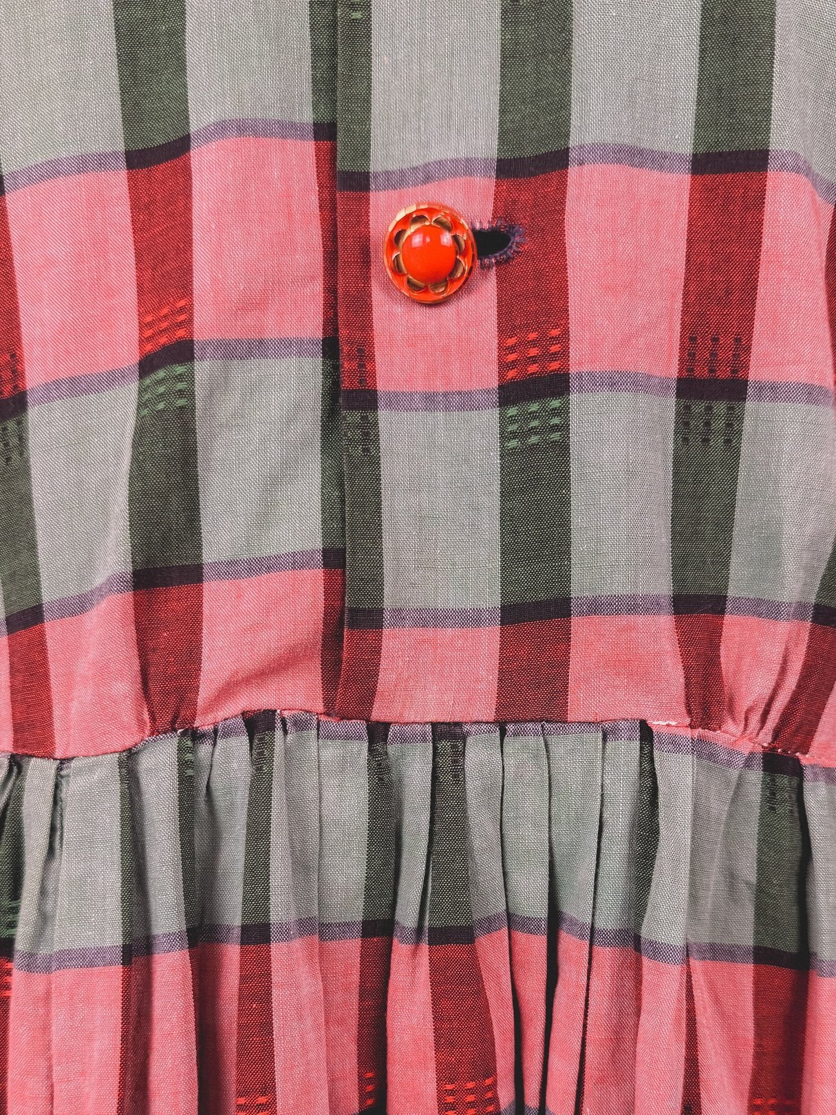 Red and green plaid fabric with a gathered waist and a red button detail, reflecting a 1950s women's holiday dress style.