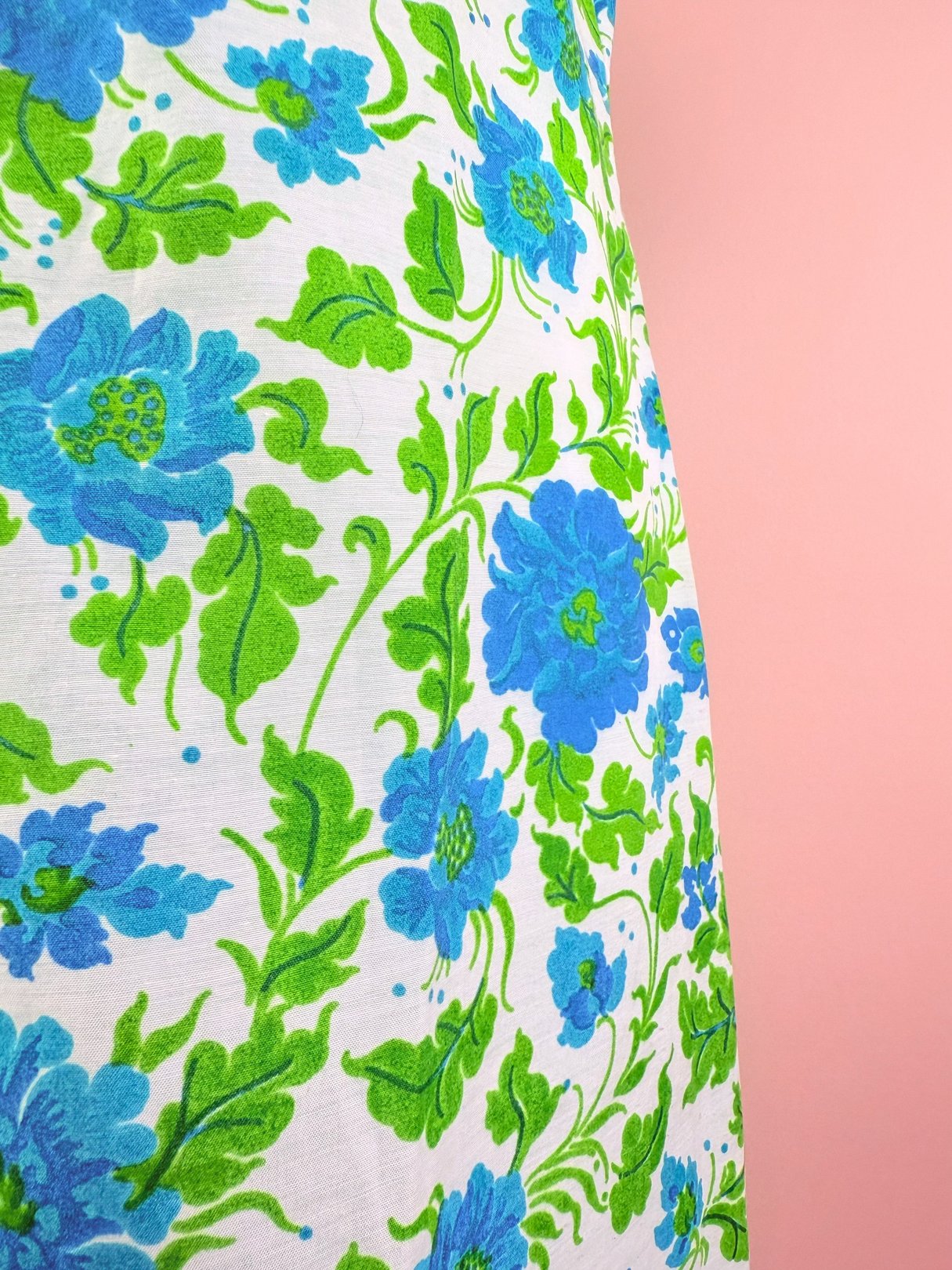 The Blue Flower River Dress features a white fabric adorned with vibrant blue and green floral patterns, characteristic of 1960s design.