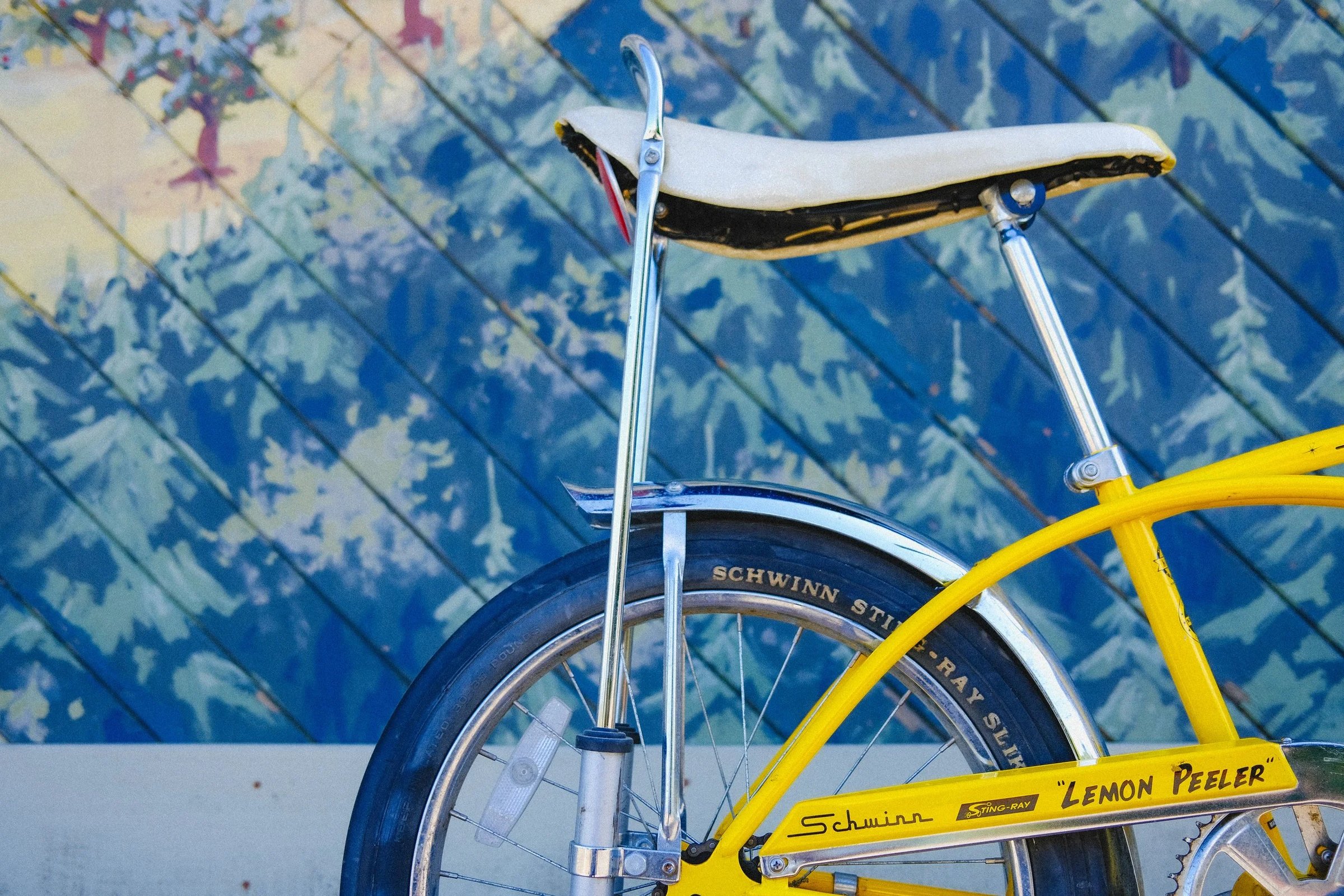 A close-up of a yellow Schwinn Lemon Peeler bicycle with a distinctive seat against a colorful mural background.