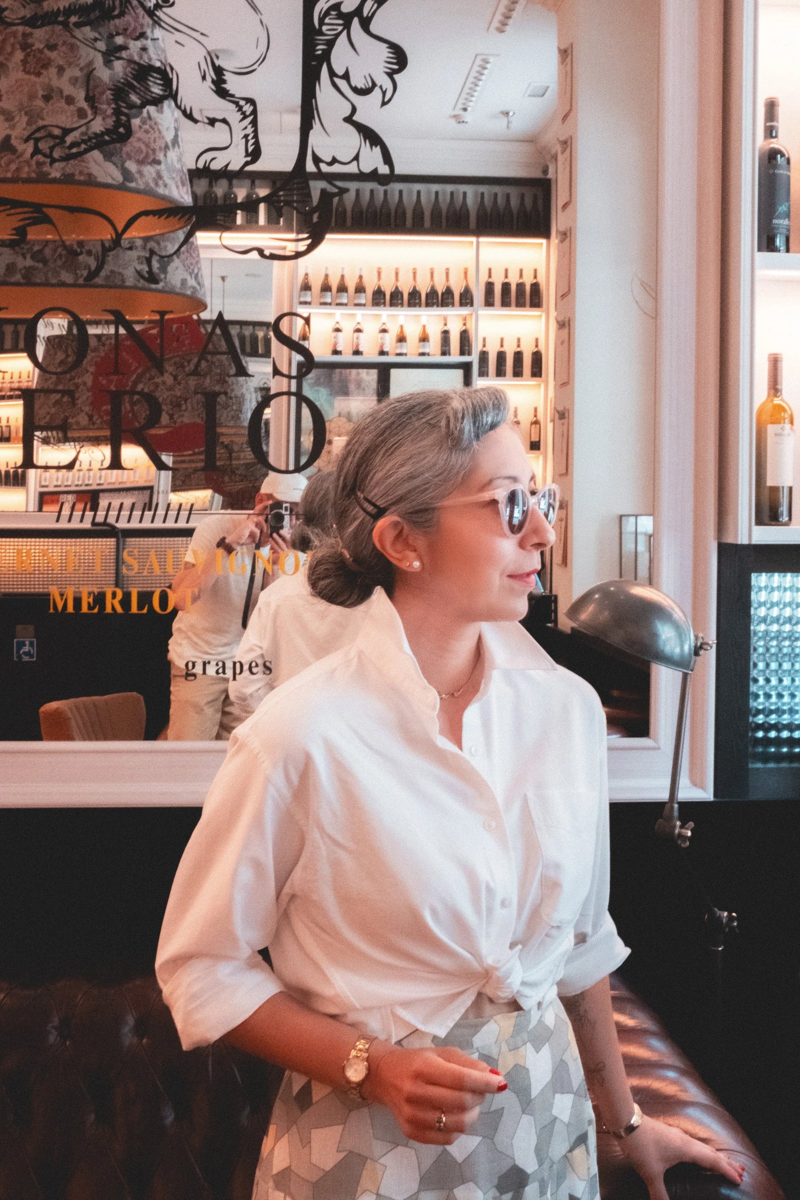 A woman with gray hair and sunglasses stands beside a wine display, wearing a tied white shirt and patterned skirt.