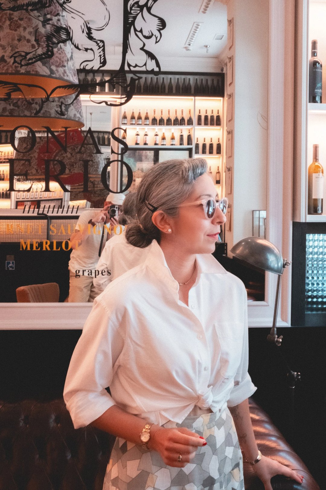 A woman with gray hair and sunglasses stands beside a wine display, wearing a tied white shirt and patterned skirt.