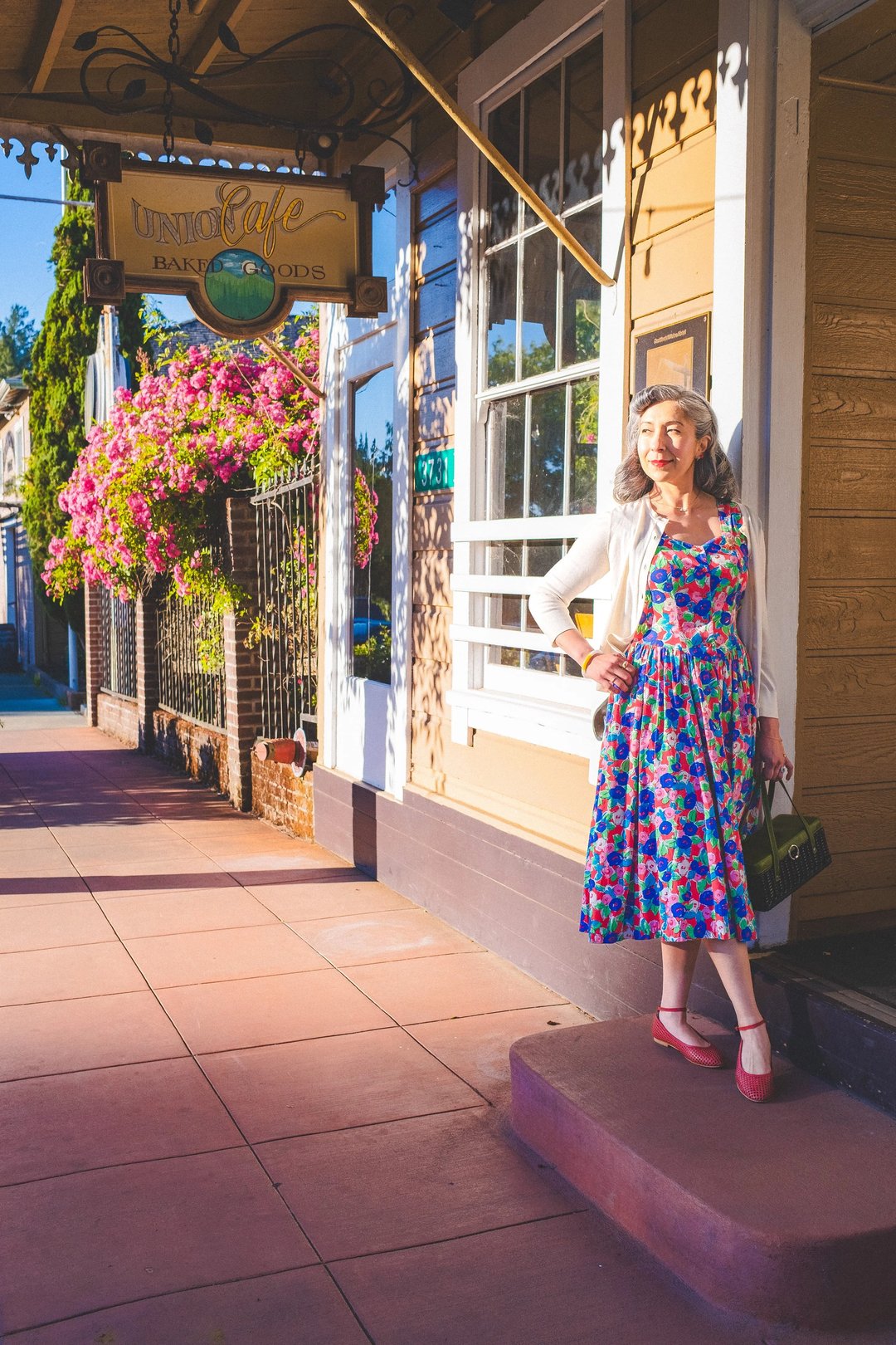 A woman in a colorful floral dress and cardigan stands on a sidewalk near a bakery, framed by blooming flowers and warm sunlight.