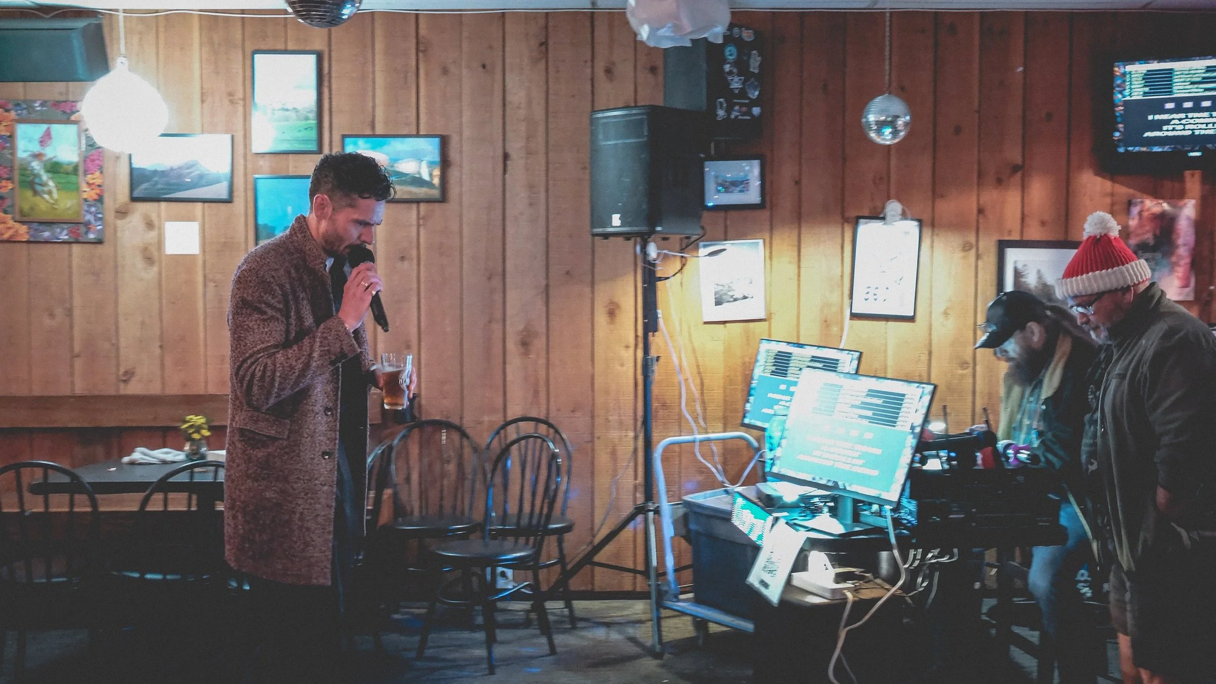 A performer sings into a microphone while standing in a rustic bar with wooden paneling and sound equipment visible.