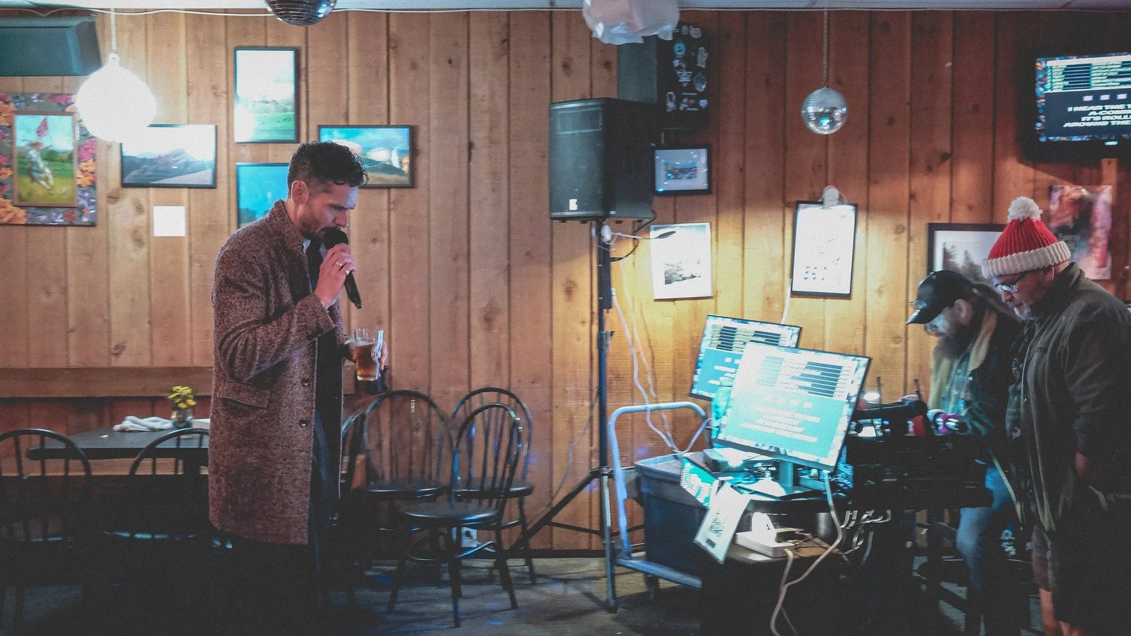 A performer sings into a microphone while standing in a rustic bar with wooden paneling and sound equipment visible.