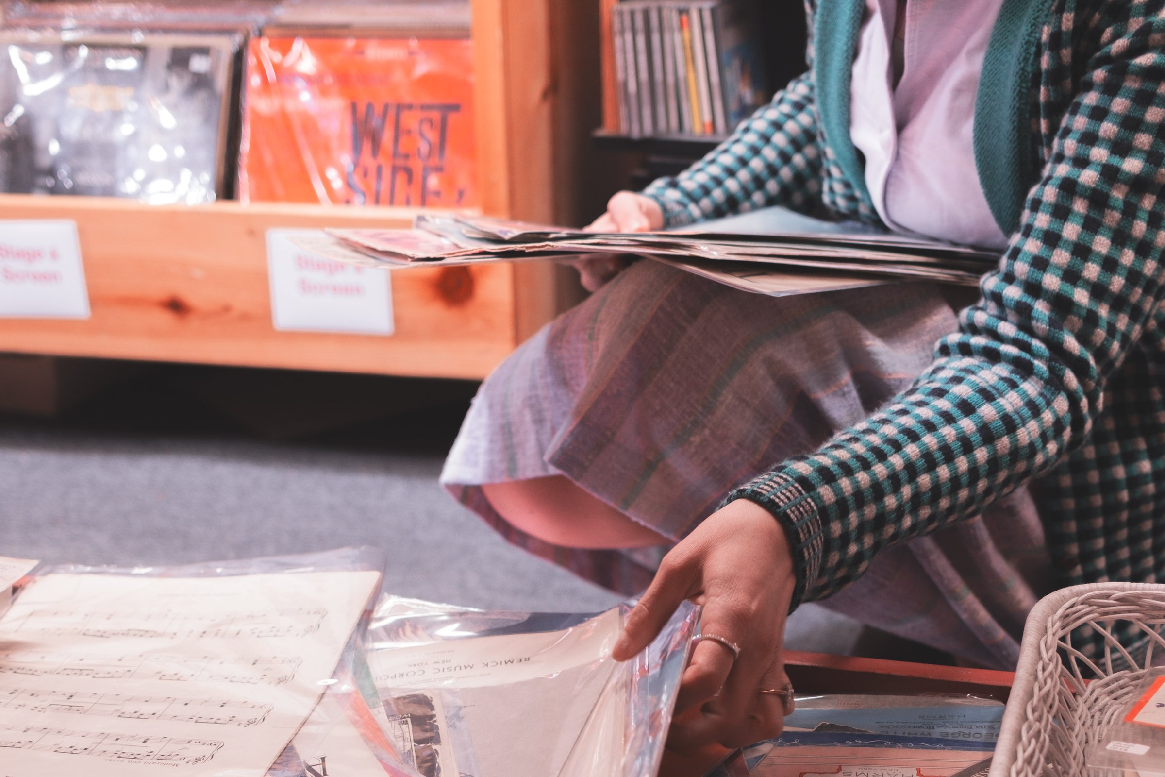 Sheet Music Stories gallery image: A person kneels on the floor, sorting through sheet music placed in protective sleeves, surrounded by shelves of music records.
