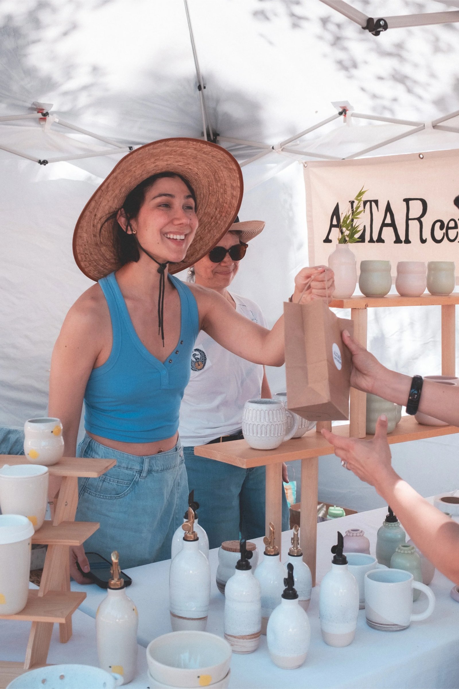 A vendor at an outdoor market hands a small paper bag to a customer while standing behind a display of pottery.