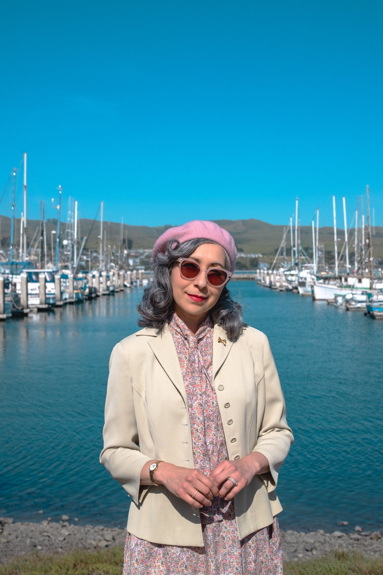 The Road to Bodega Bay gallery image: A woman in a pastel beret and sunglasses stands near a marina with sailboats and clear blue skies in the background.