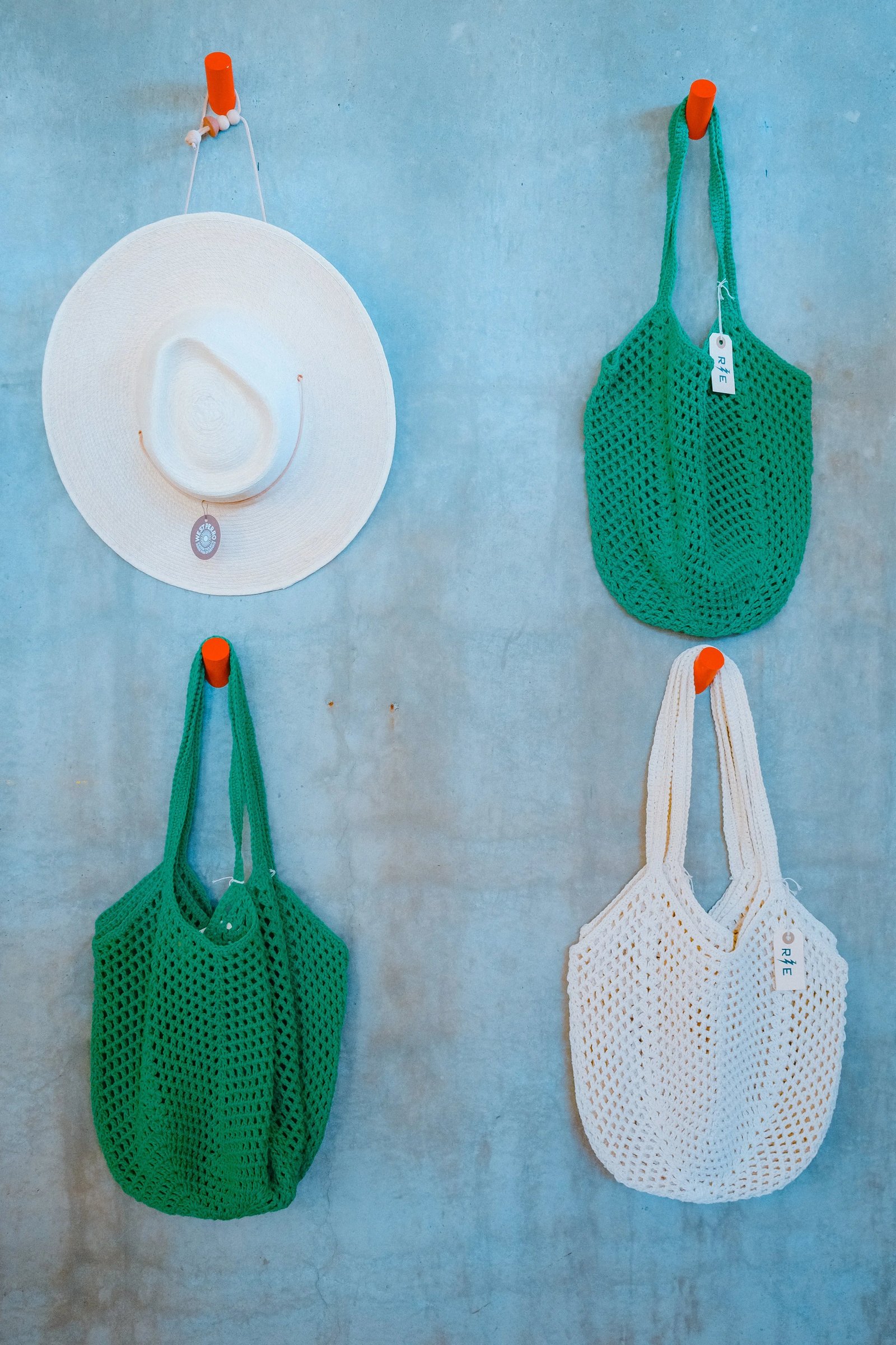 Two green mesh tote bags and a white mesh tote bag hanging on hooks next to a white sun hat against a concrete wall.