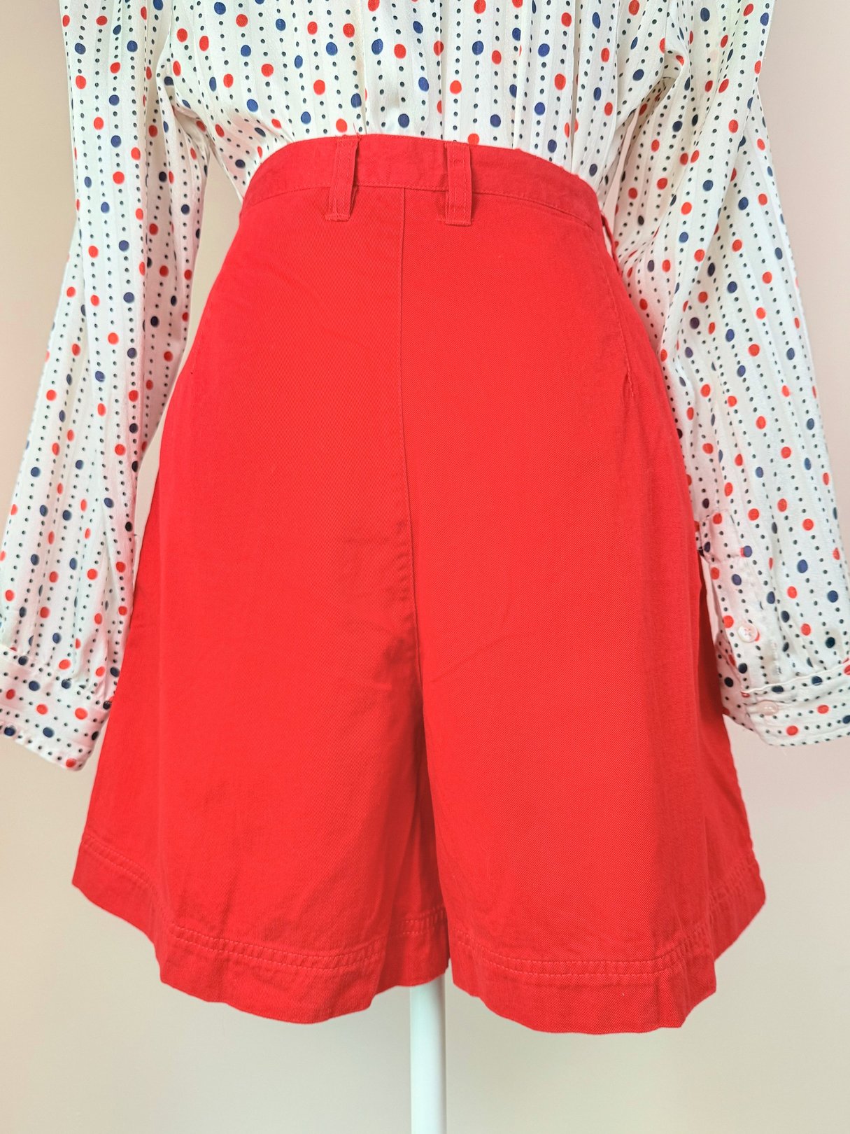 High-waisted red shorts with a relaxed fit, styled with a long-sleeved polka dot shirt, reflecting 1980s fashion.