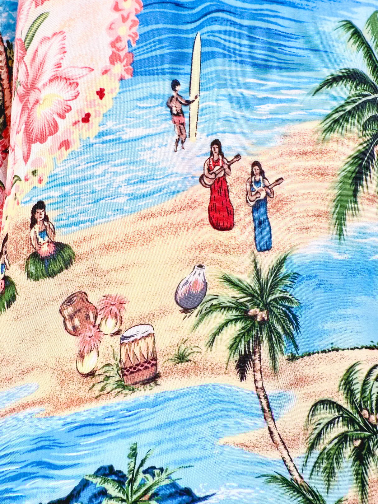 The Aloha Darling Top features a vibrant blue fabric with a 1960s tropical floral print, depicting surfers, musicians, and palm trees.