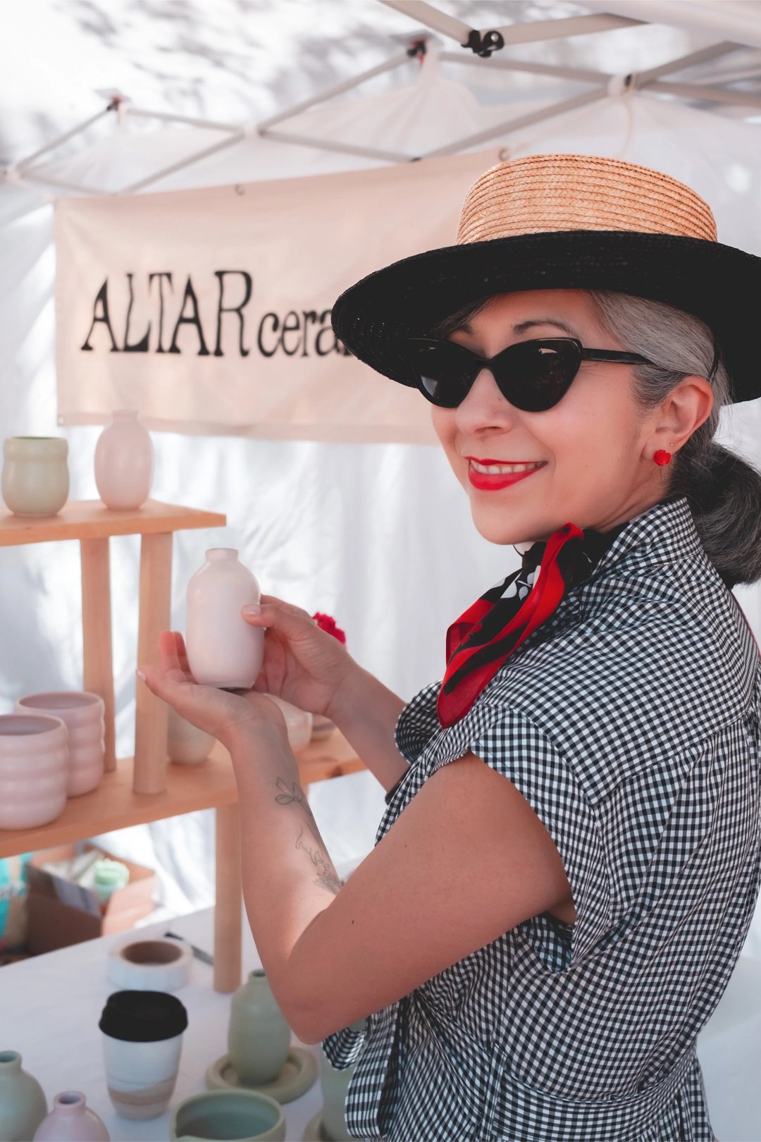 A woman wearing a straw hat and sunglasses smiles while holding a white ceramic vase at a market stall displaying various pottery items.