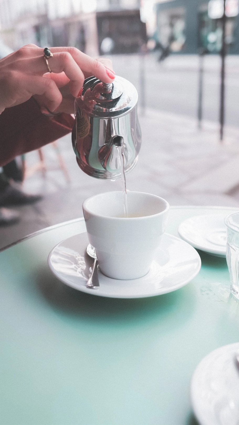 A hand pours hot water from a silver teapot into a white cup on a table.