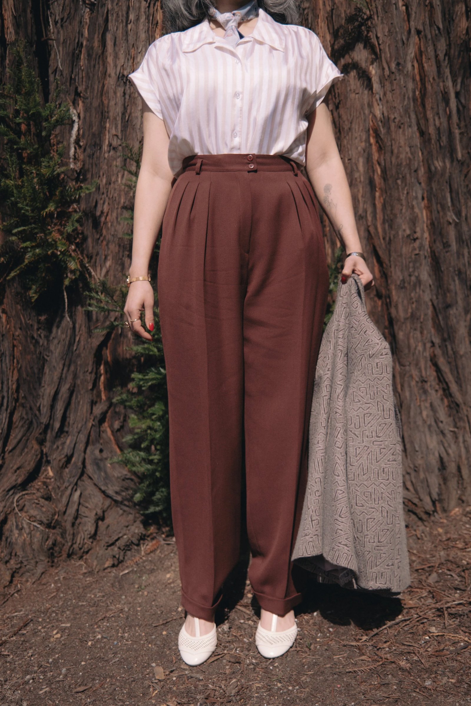 A woman stands in a natural setting, wearing a white top and brown high-waisted trousers, holding a light jacket.