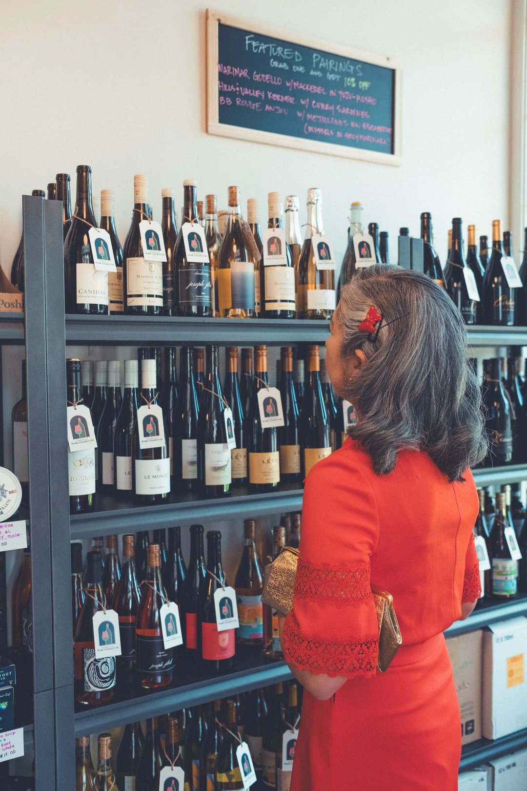 A woman in a red dress examines a selection of wine bottles on a shelf with label tags.