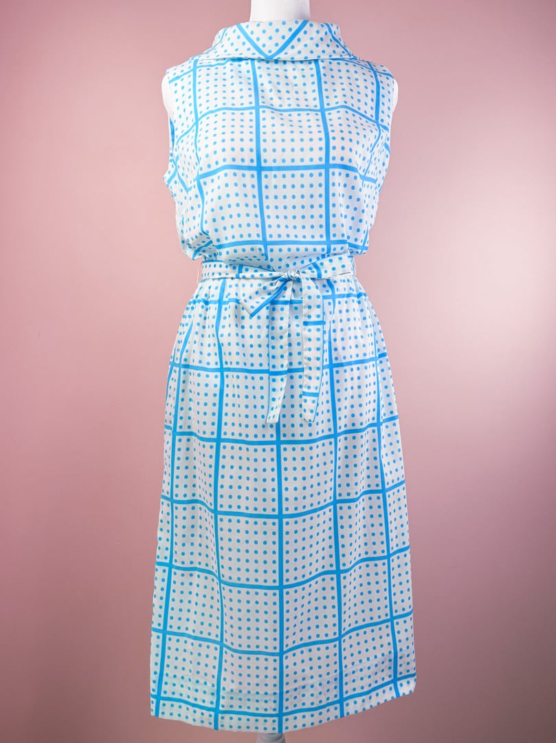 A sleeveless white dress with a blue grid pattern and a tied waist, reflecting a 1970s style.