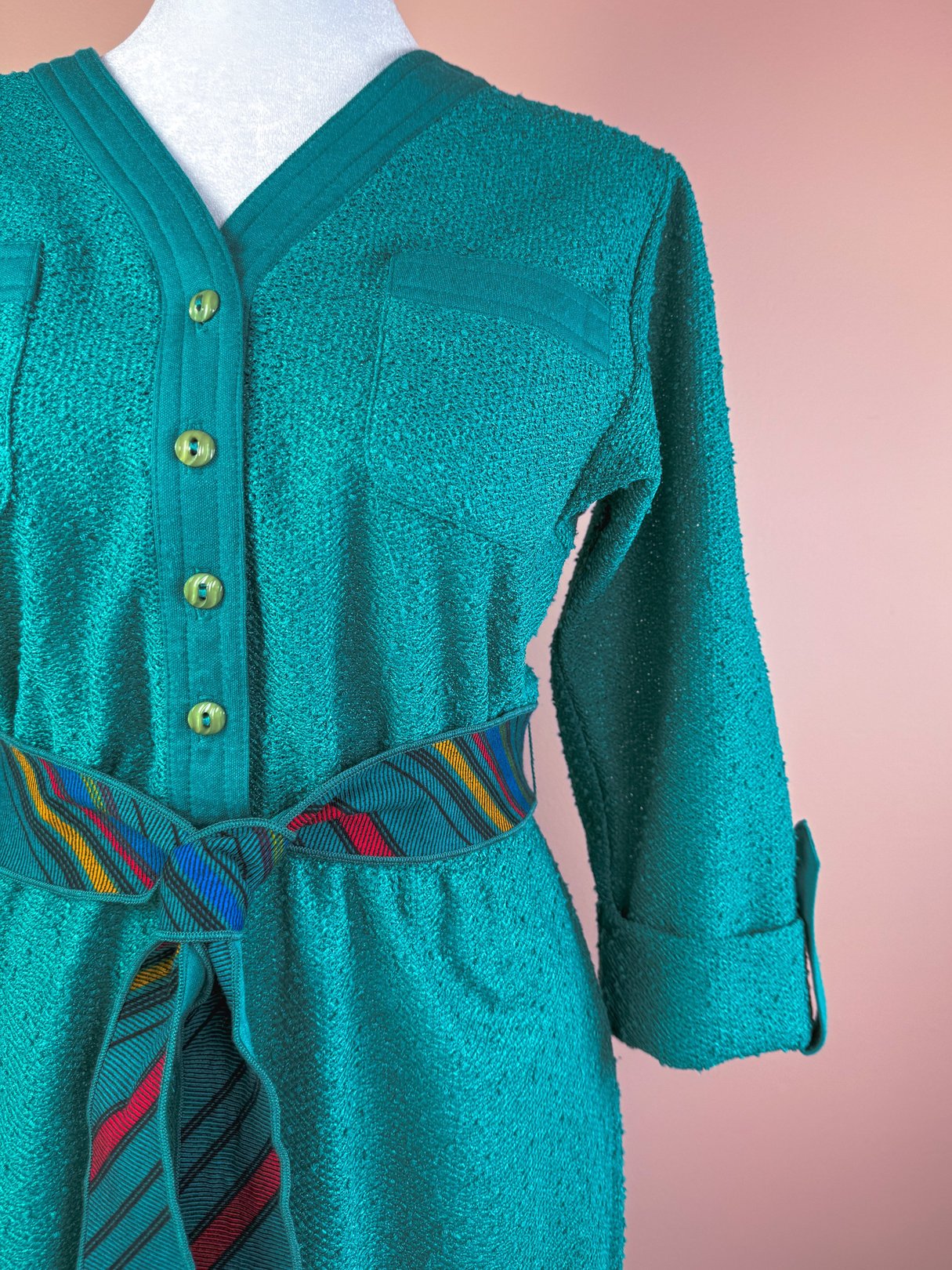 Teal dress with a V-neck, buttoned front, a chest pocket, and a multi-colored striped belt, featuring 1980s-inspired textured fabric.