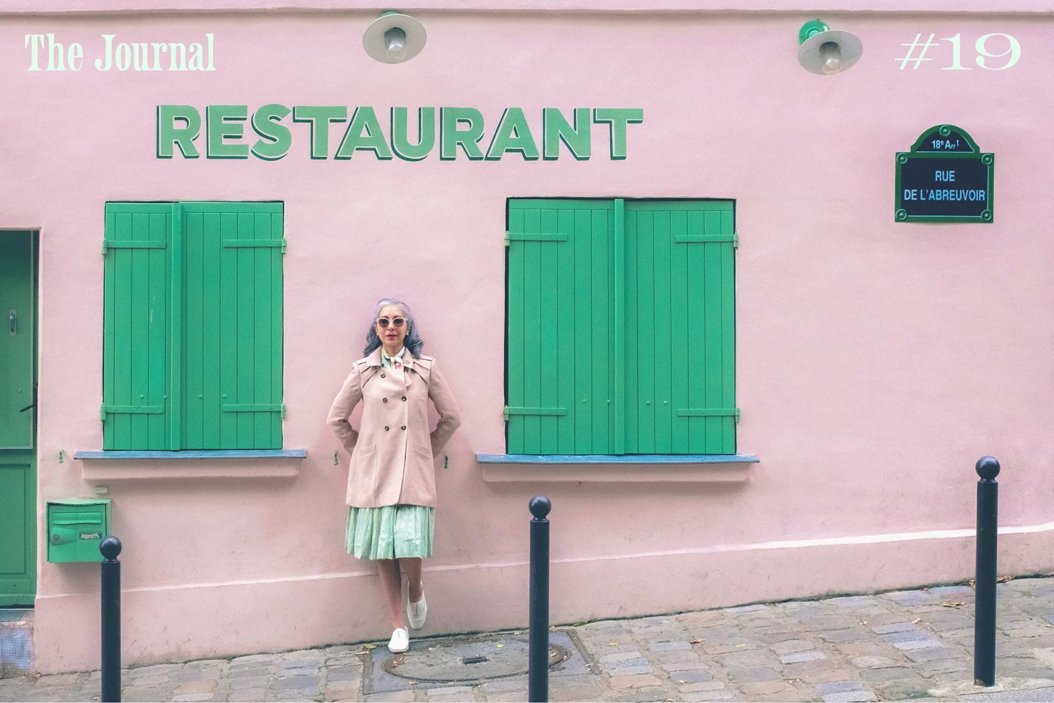 A woman in a beige coat and sunglasses stands in front of a pink restaurant wall with green shutters and signage.