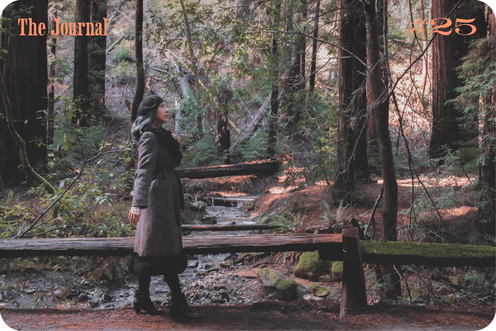A woman in a gray coat stands by a wooden bridge in a lush forest, with a creek running in the background.