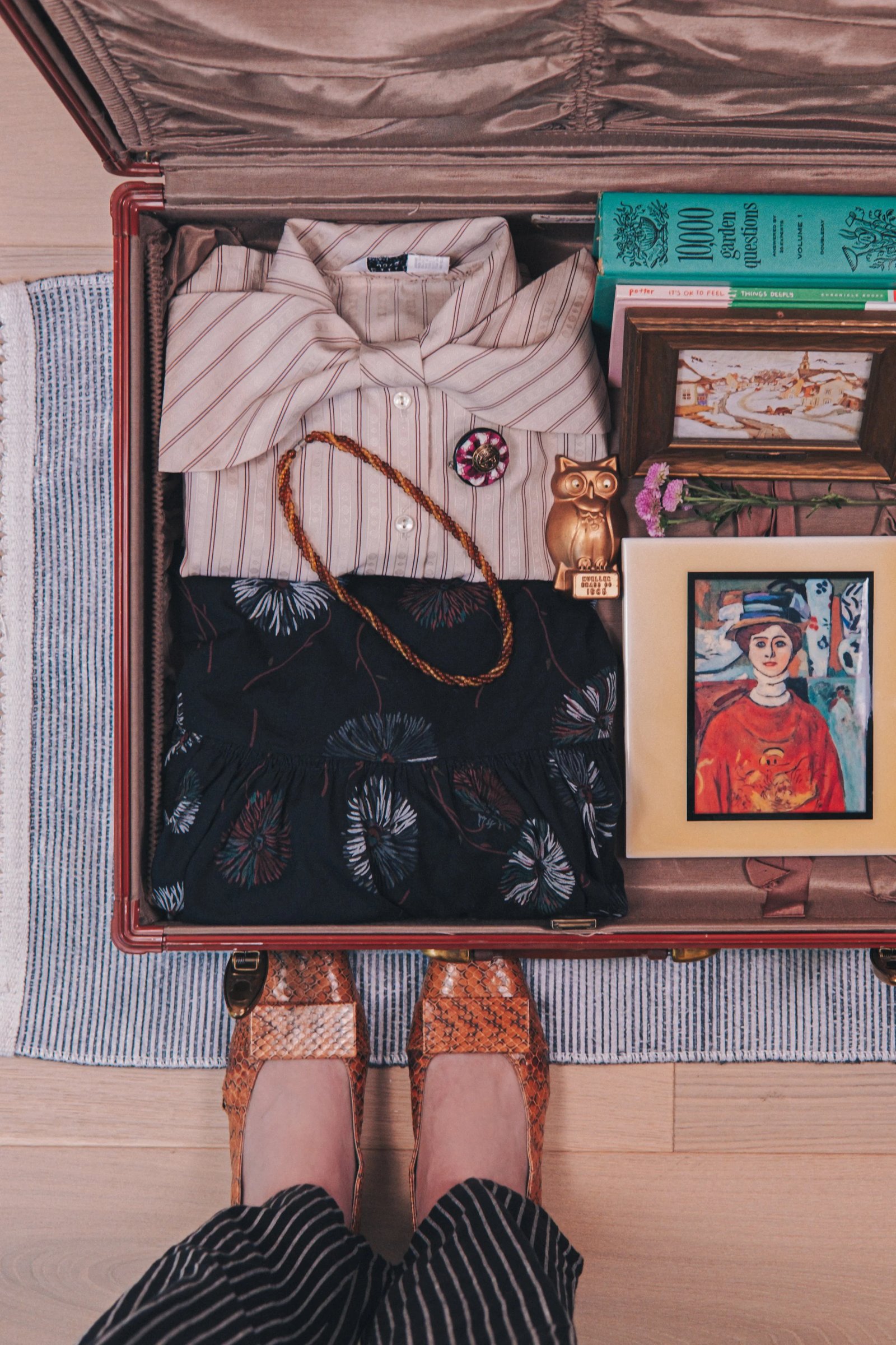 A vintage suitcase contains neatly folded clothing, accessories, framed artwork, and a decorative owl figurine, with a pair of shoes in the foreground.